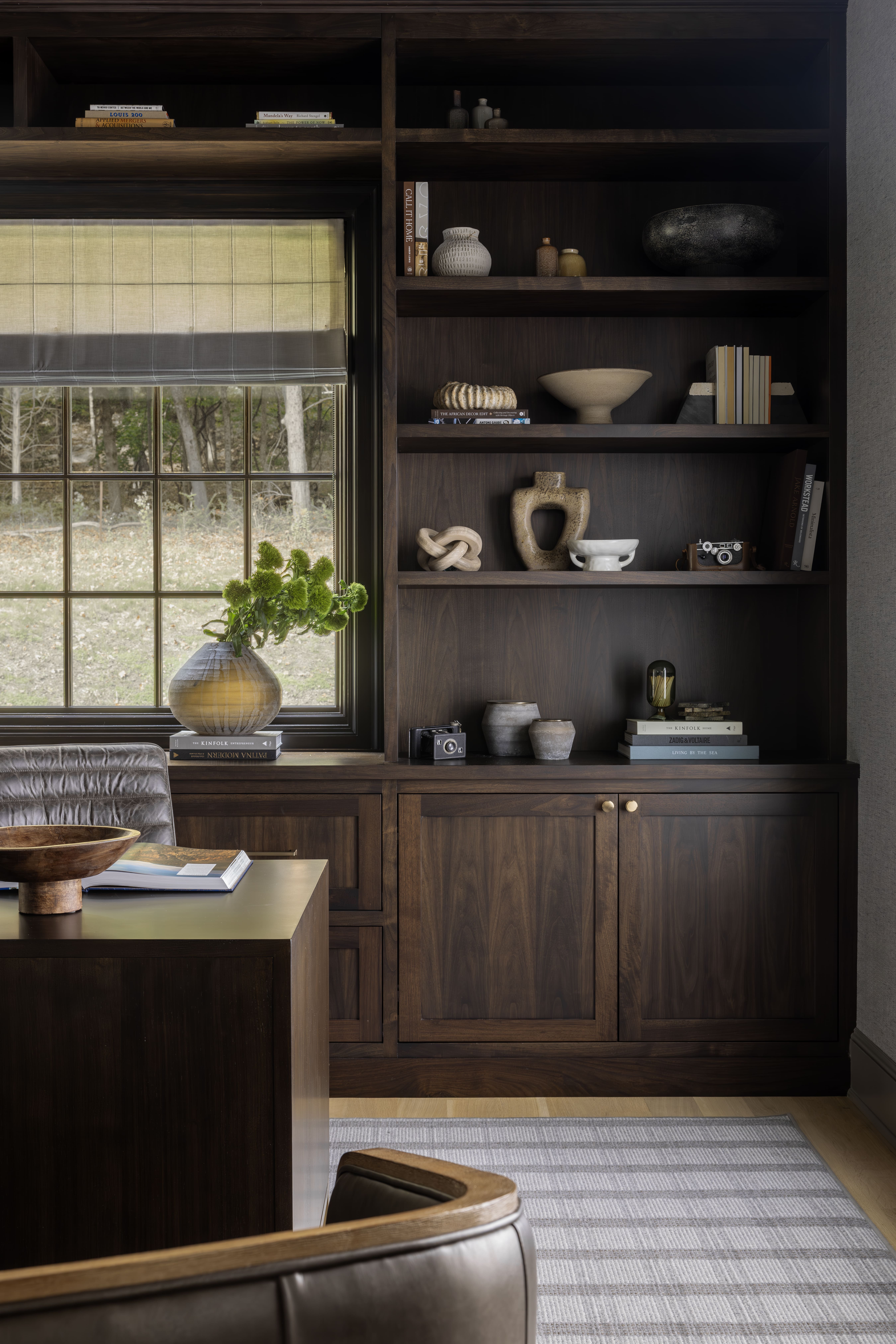 Dark wood built-in shelving with decorative pottery and books beside a large window with a striped shade