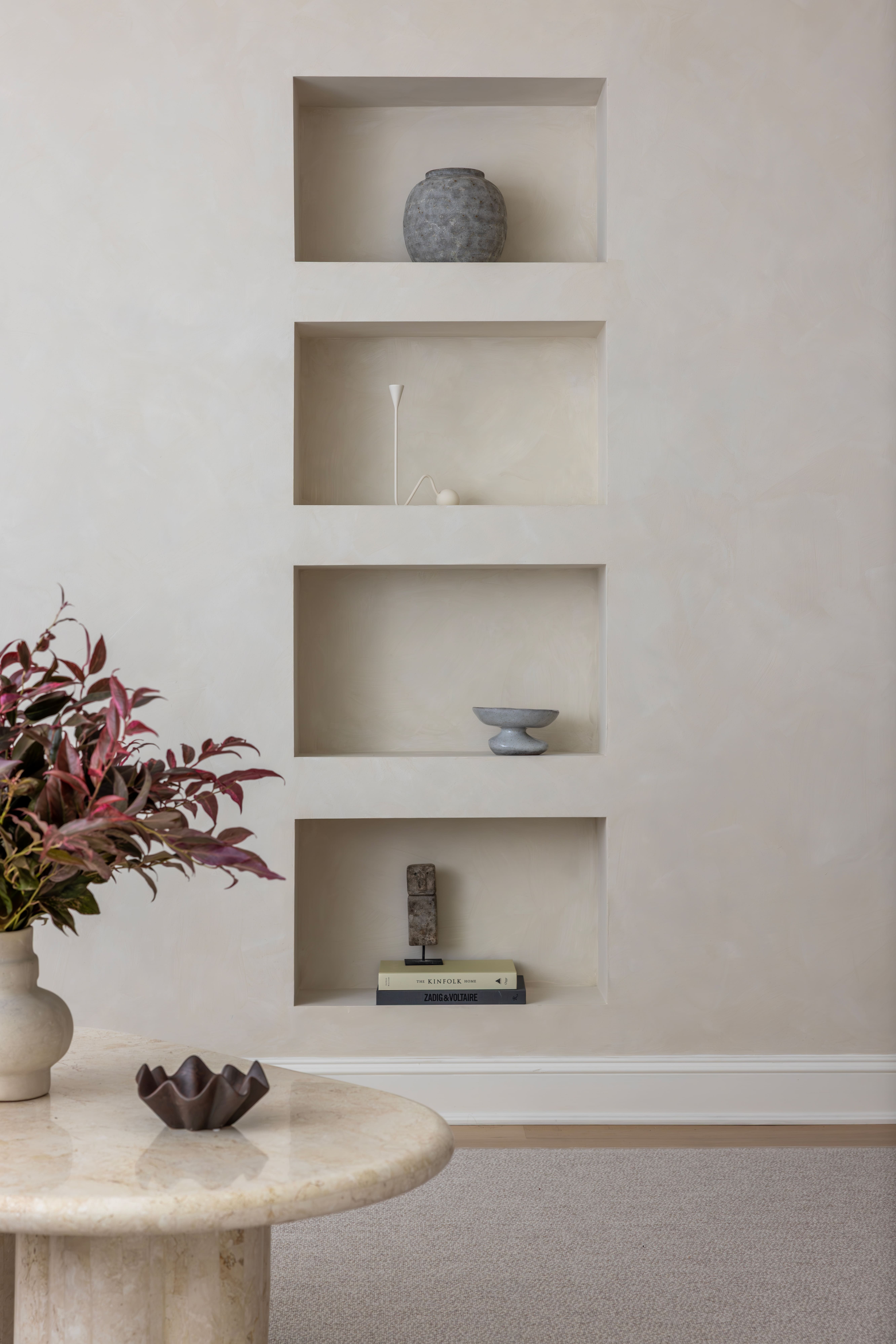 Neutral built-in wall shelving with four recessed niches displaying minimalist decor: a gray vase on the top shelf, a white modern lamp on the second, a small sculpture on the third, and a stack of books with a tiny figurine on the bottom shelf; a vase with burgundy leaves sits to the left.