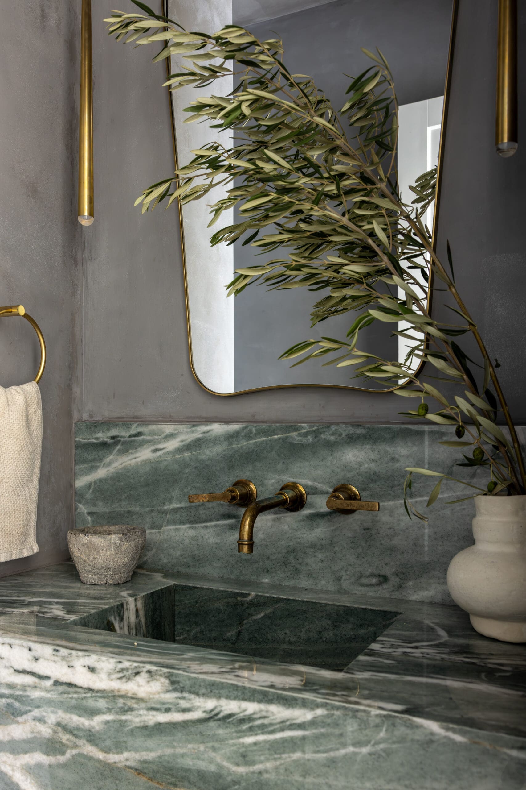 Brass bathroom fixtures with a large mirror and olive branch resting on a gray marble sink wall.