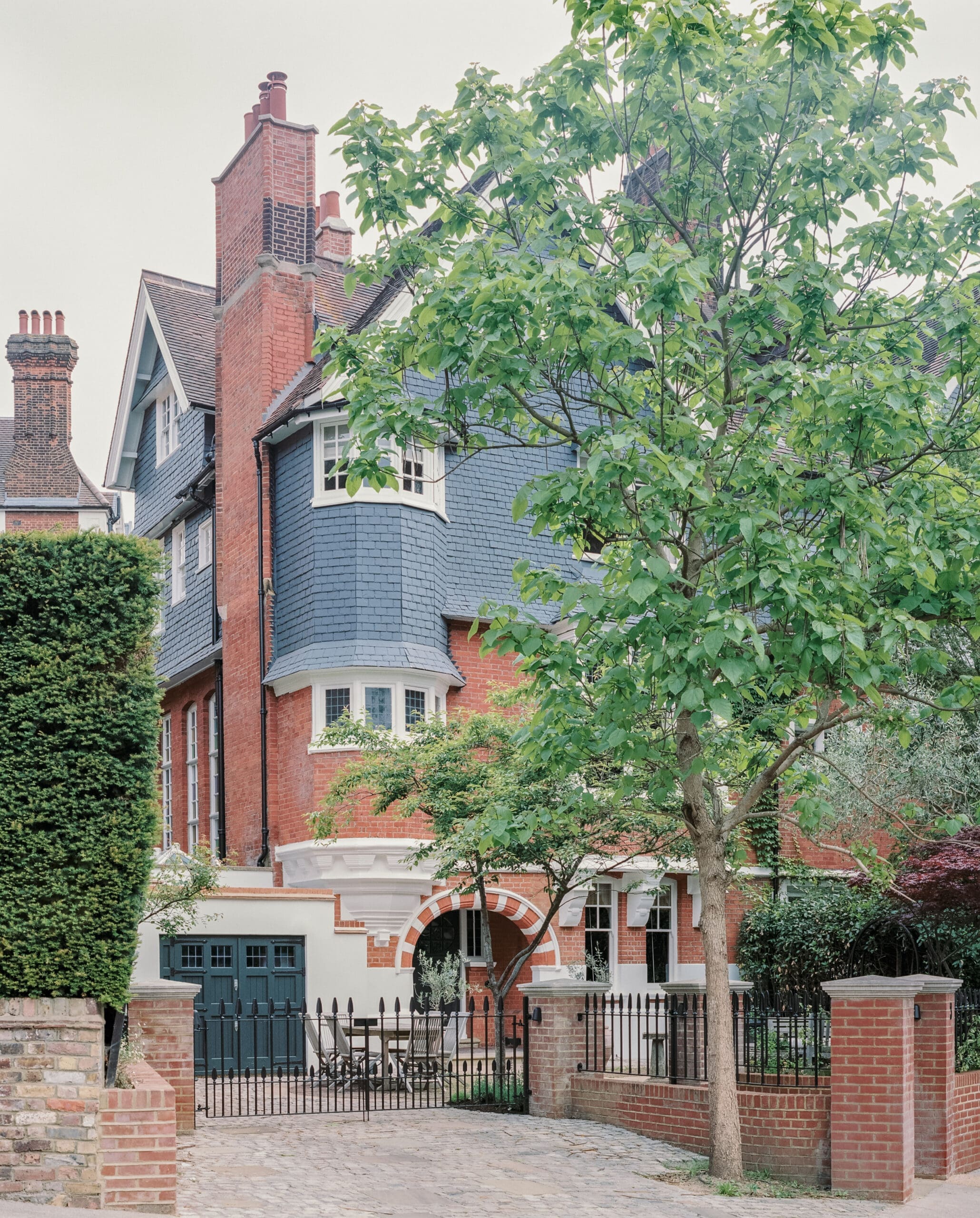 Red brick townhouse with blue slate roof behind a black iron fence and a leafy tree in front