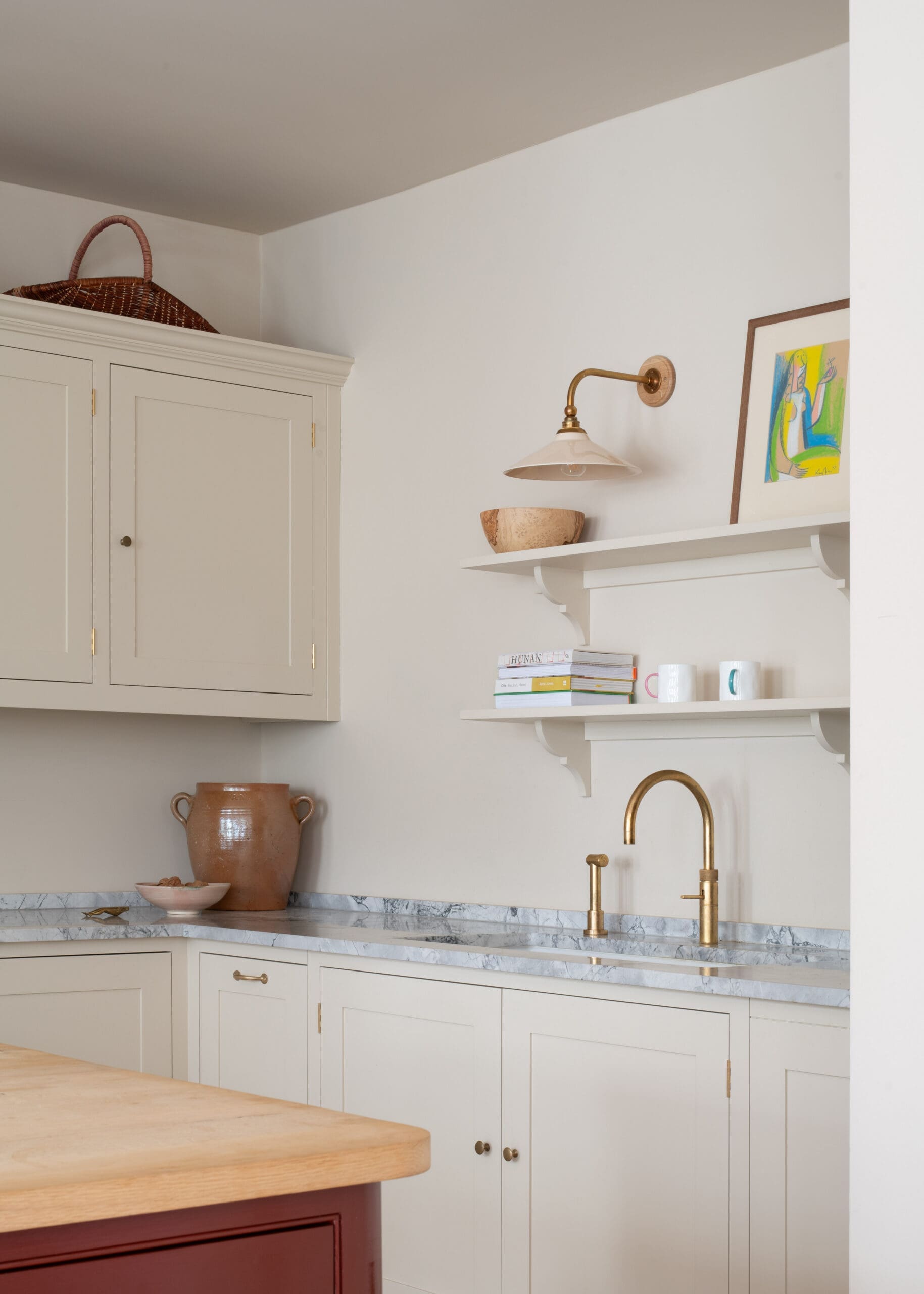 Cream kitchen cabinets with a marble countertop, brass faucet, and a wall shelf holding bowls and a colorful framed artwork.