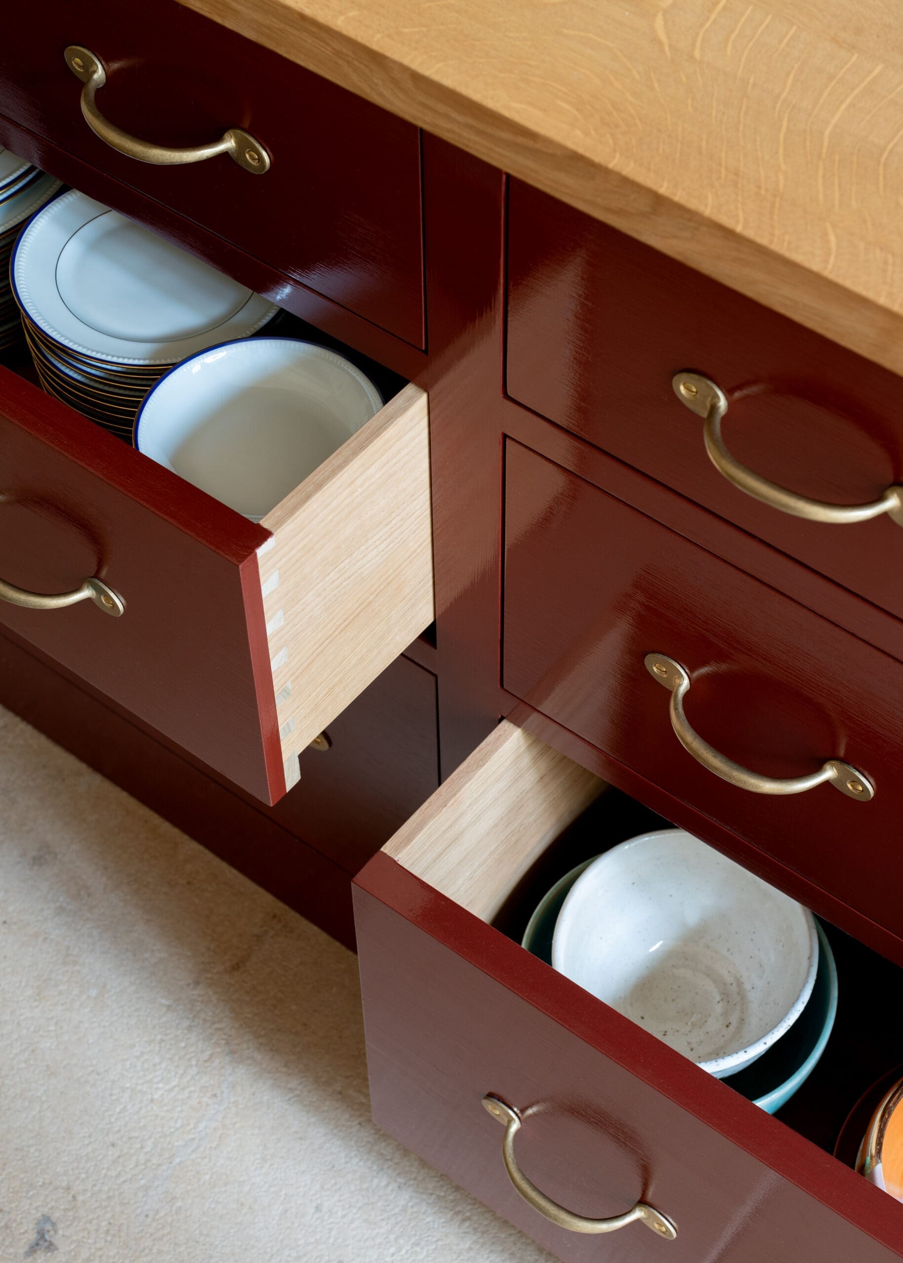 Open kitchen drawer revealing stacked white plates with blue rims and bowls inside a dark red cabinet with brass handles.