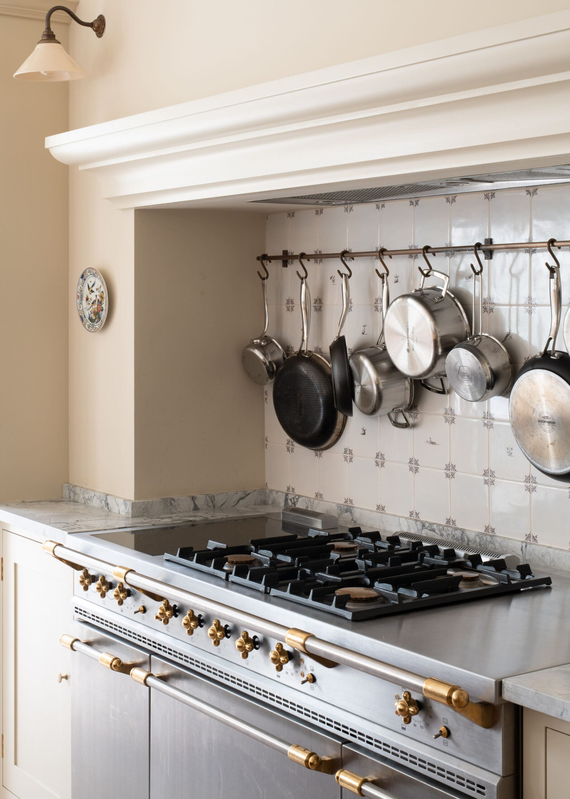 Stainless steel kitchen range with multiple pots hanging from a rack above a tiled backsplash.