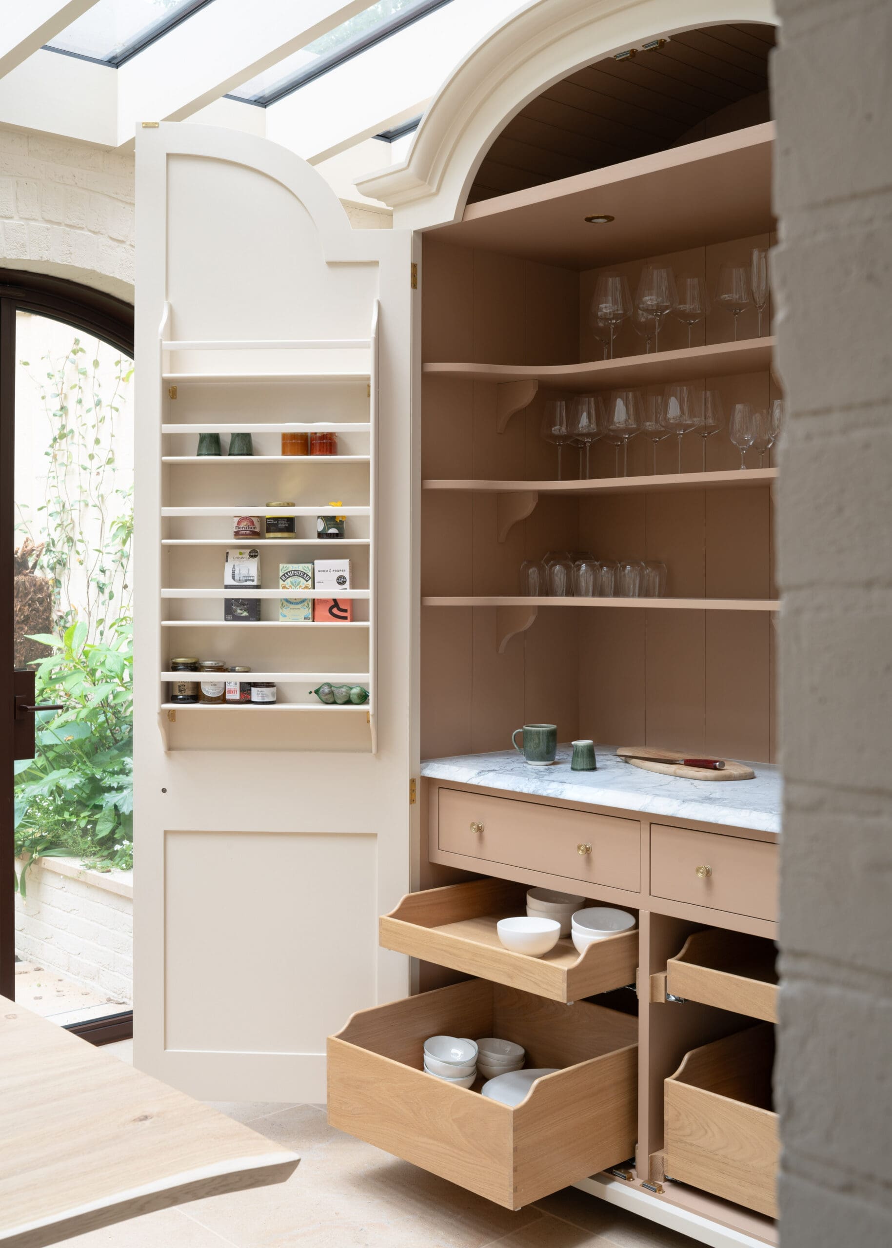 Open kitchen pantry with shelves full of glassware and jars, and pull-out wooden drawers containing bowls on a marble counter.
