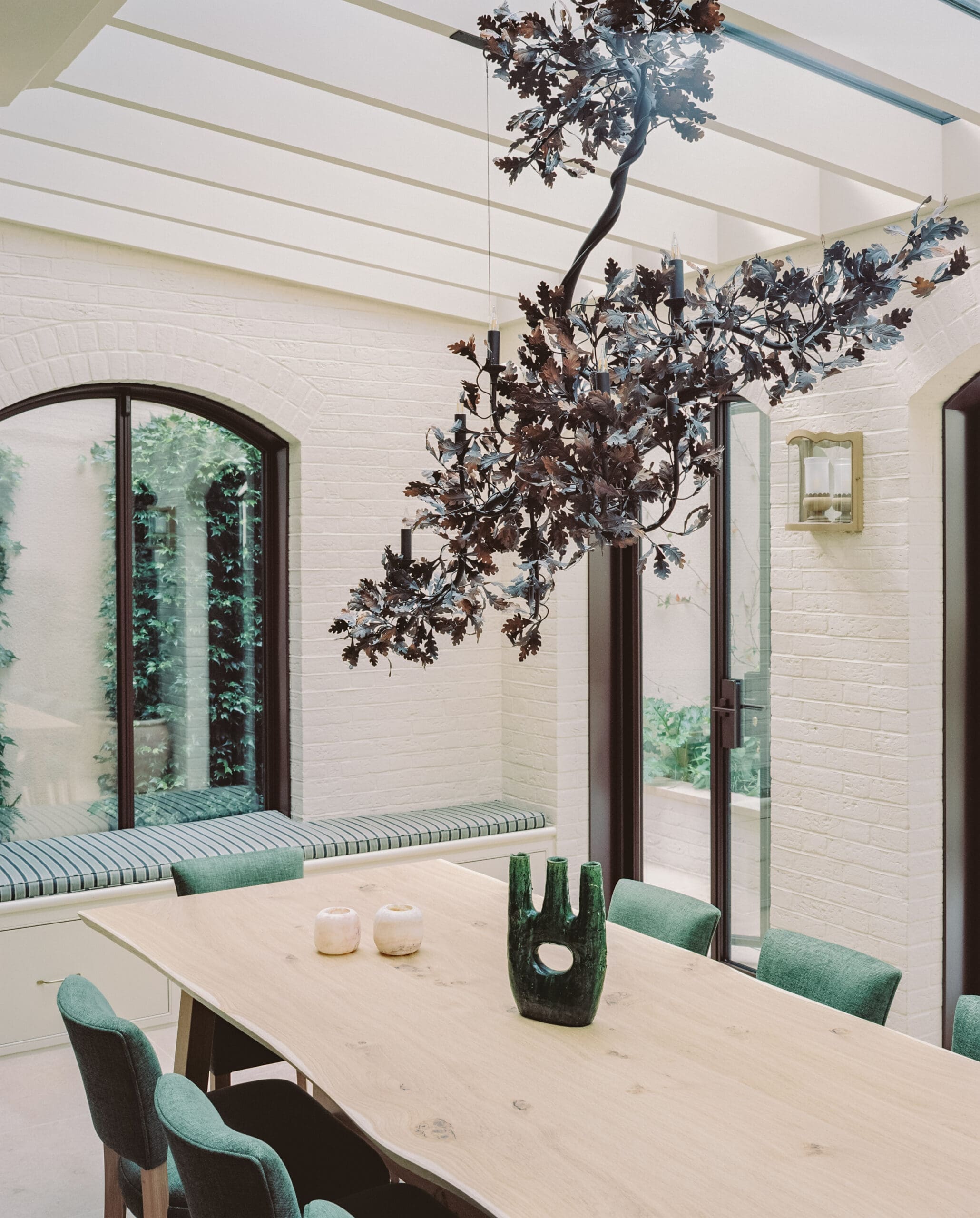 Sunlit indoor dining area with a long light wood table, green upholstered chairs, and a dark leafy chandelier centerpiece above.