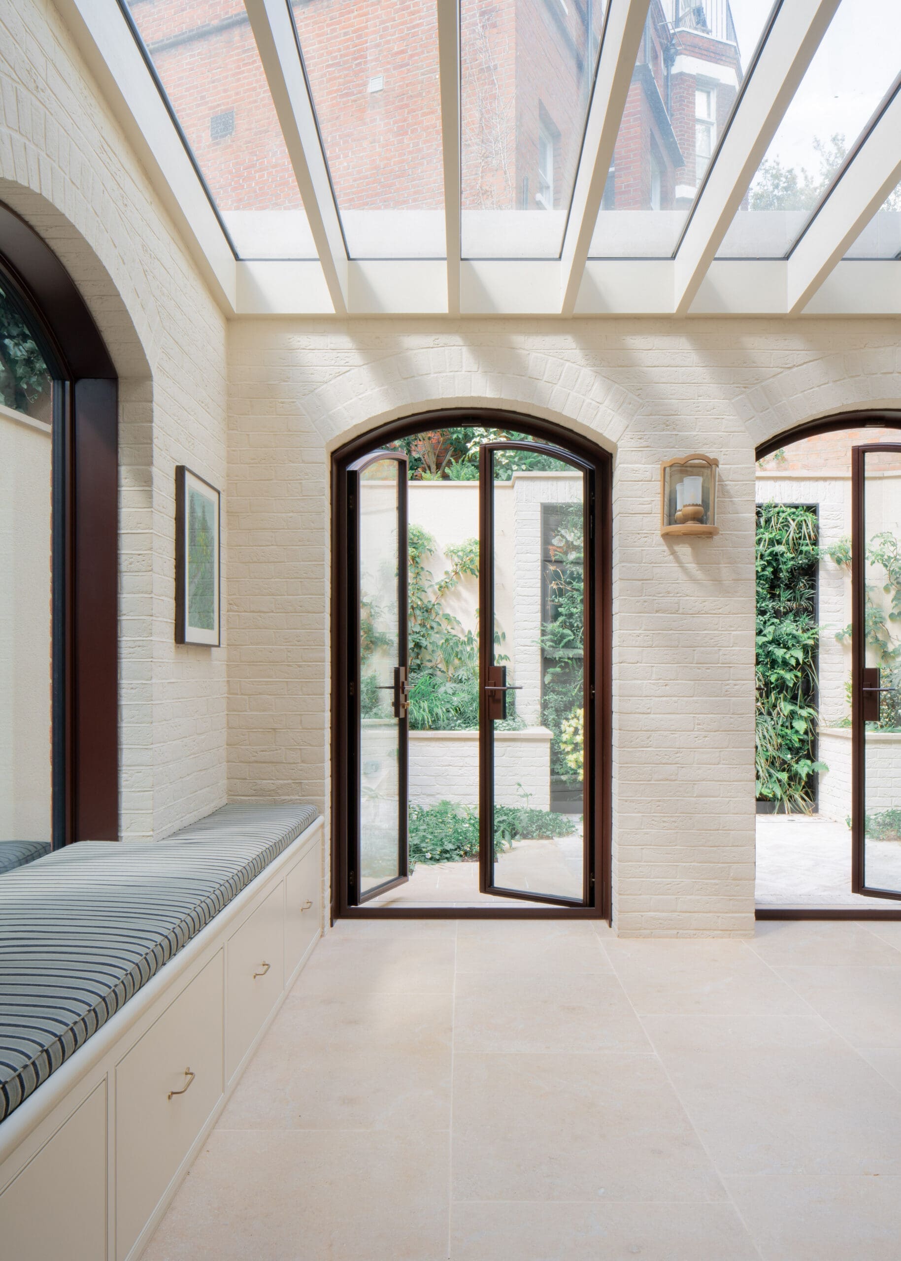 Bright hallway with arched glass doors and skylight, white brick walls, and built-in bench with striped cushion.