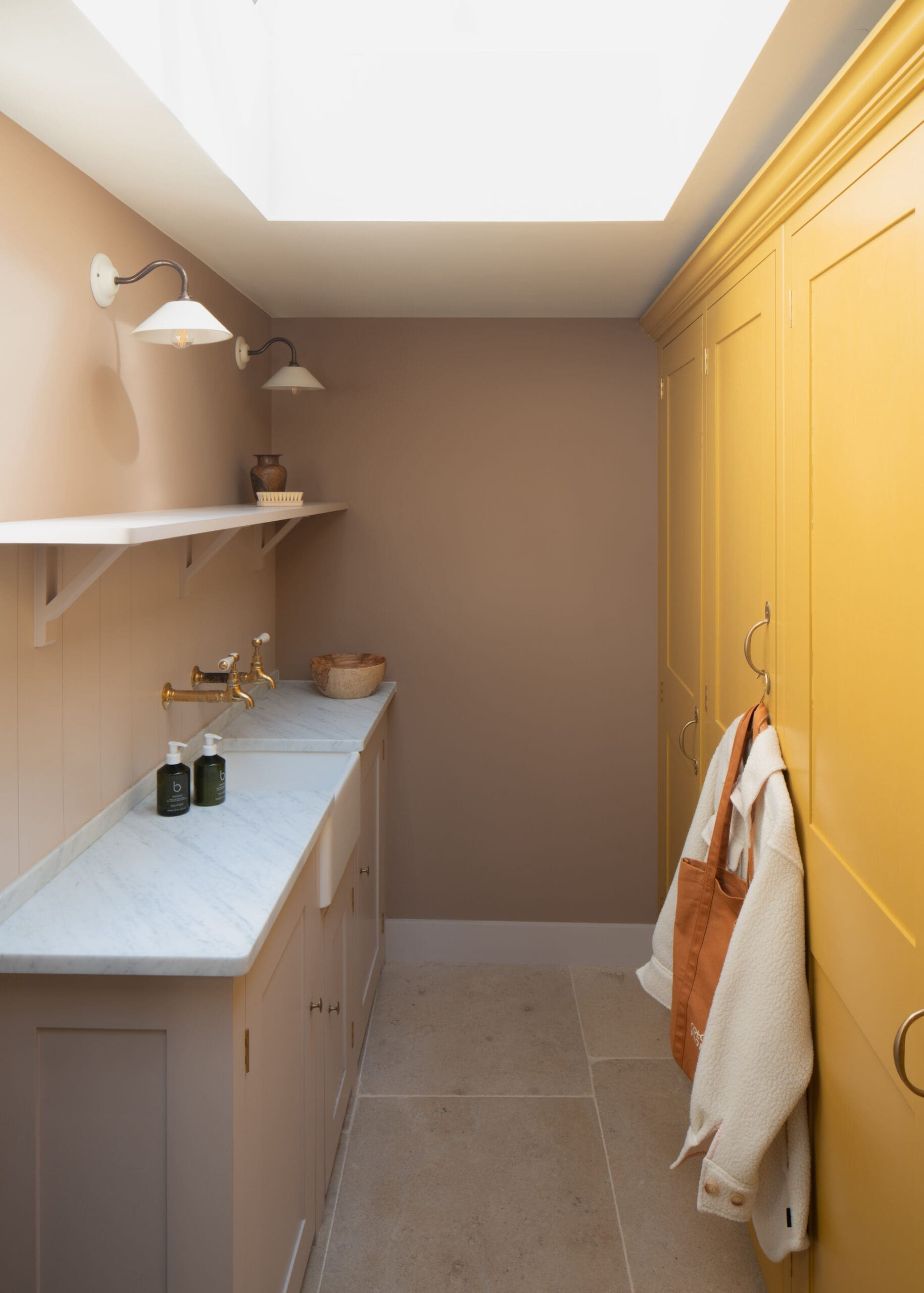 Narrow bathroom with beige walls, marble countertop, brass faucets, and two wall-mounted lamps; yellow built-in cabinets and towels on a door.