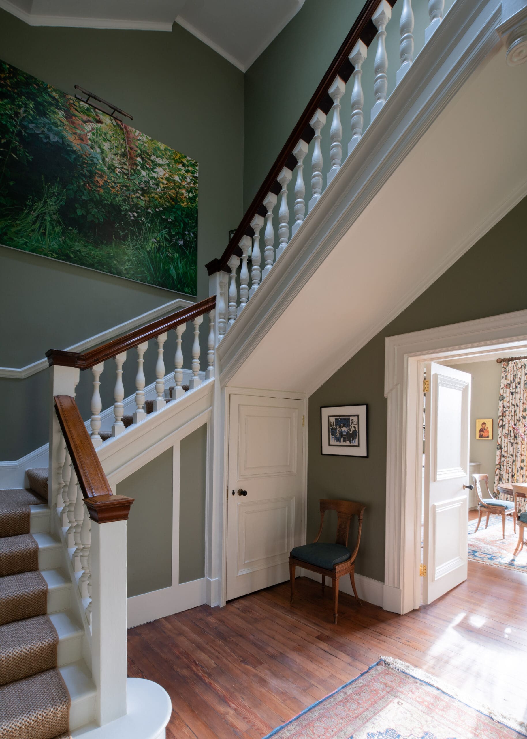 Interior stairhall with white balustrade and dark wood handrail along green walls; large greenery painting on the wall and doorway to a bright room beyond