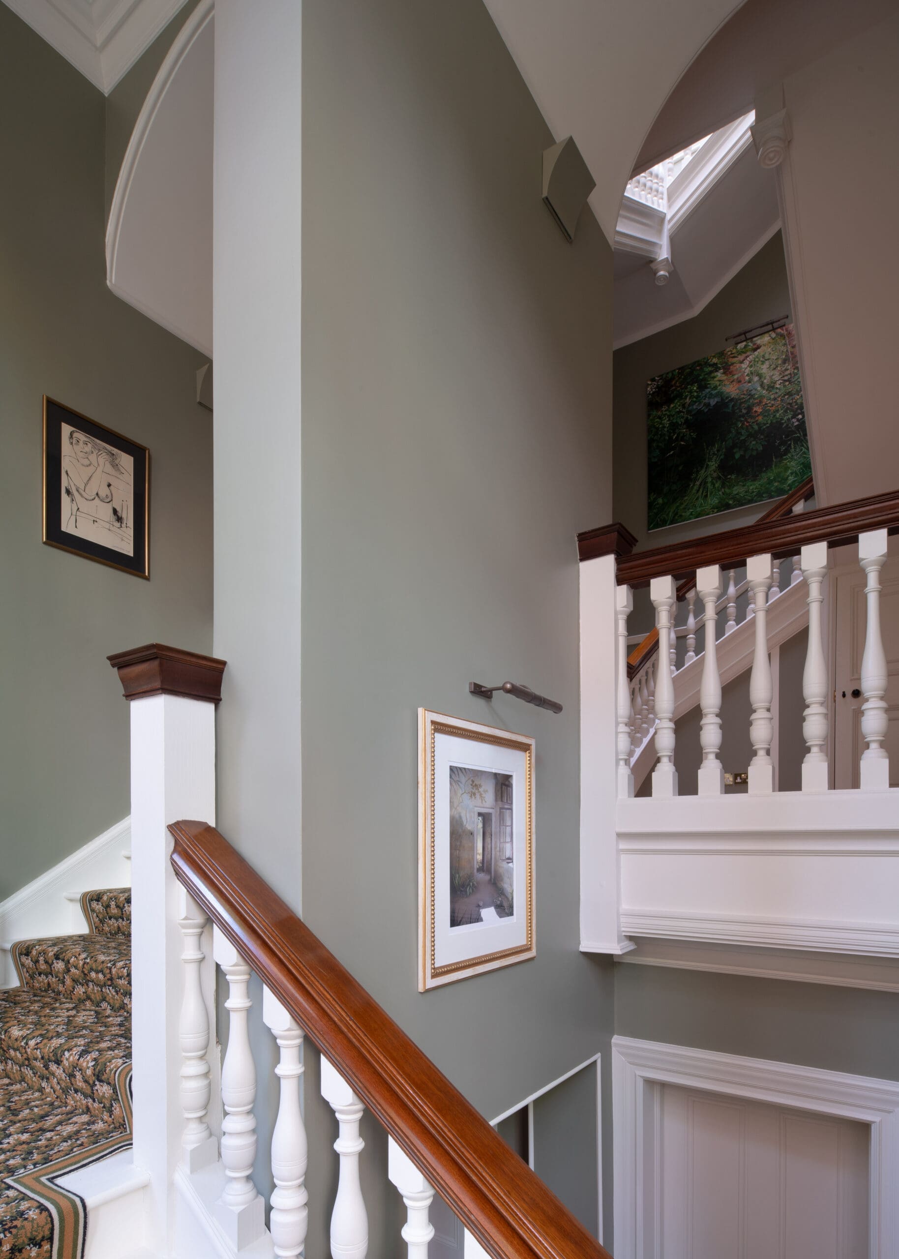 Interior staircase with a polished wooden handrail, white balusters, and green walls, framed artwork on the wall (stairwell scene).
