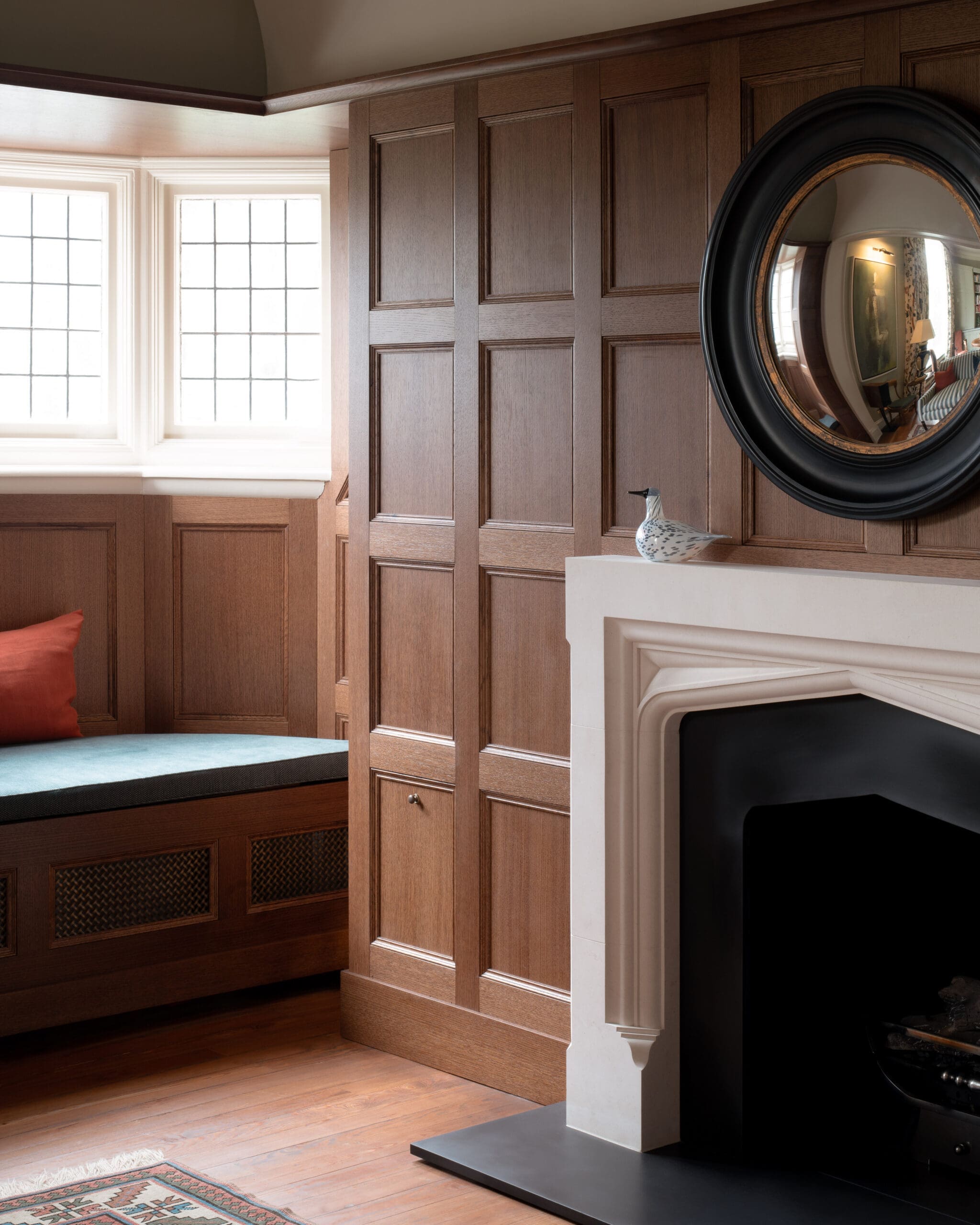 Wood-paneled wall with a large round black-framed mirror above a white fireplace mantel; a ceramic bird sits on the mantel, with sunlit windows to the left.
