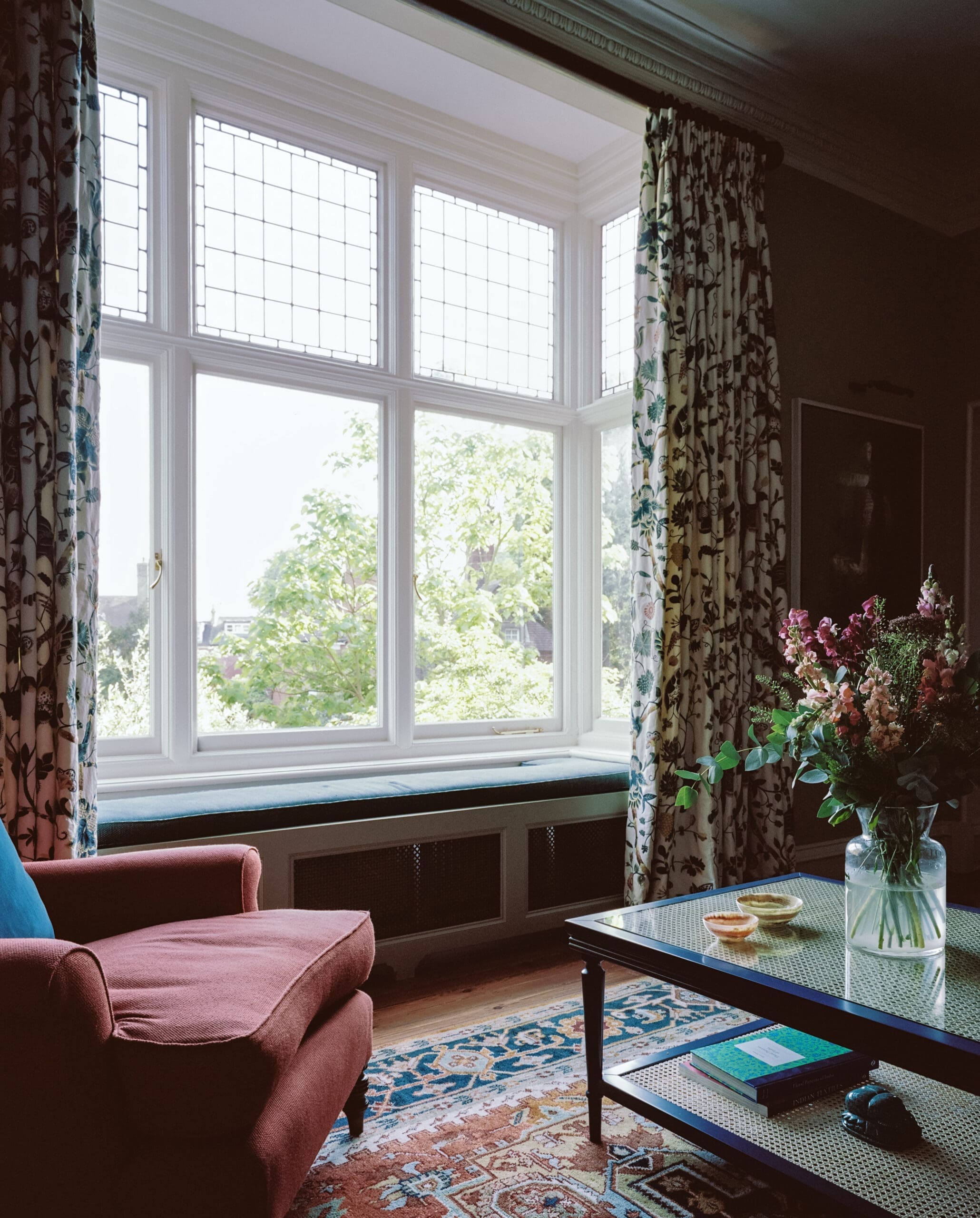 Sunlit living room with a bay window, floral curtains, and a pink sofa nearby. A glass coffee table with a vase of flowers sits in front of the window.
