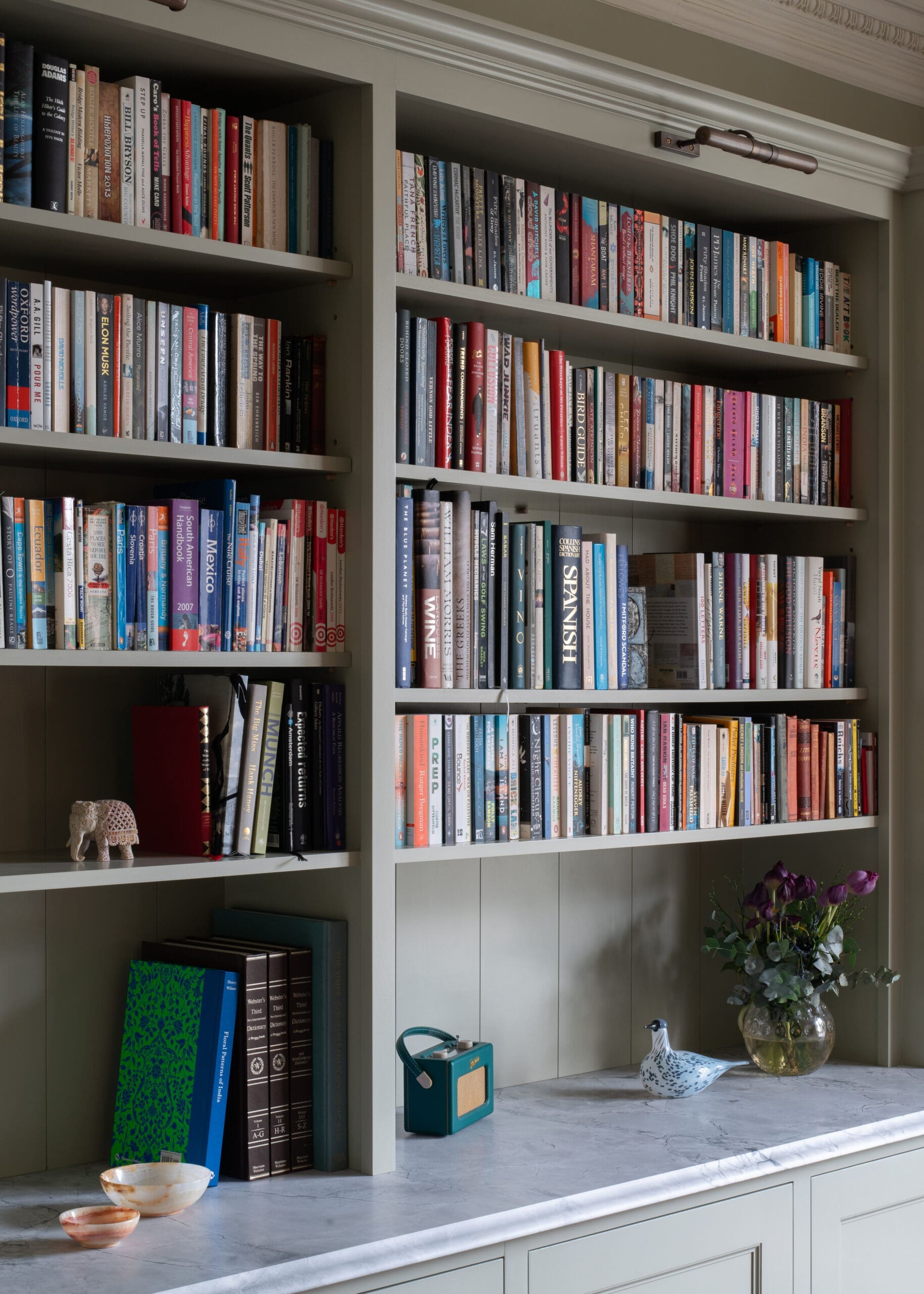 Built-in shelving filled with colorful books, plus a small elephant figurine, a teal vintage radio, white ceramic birds, and a vase of purple flowers on a marble counter.