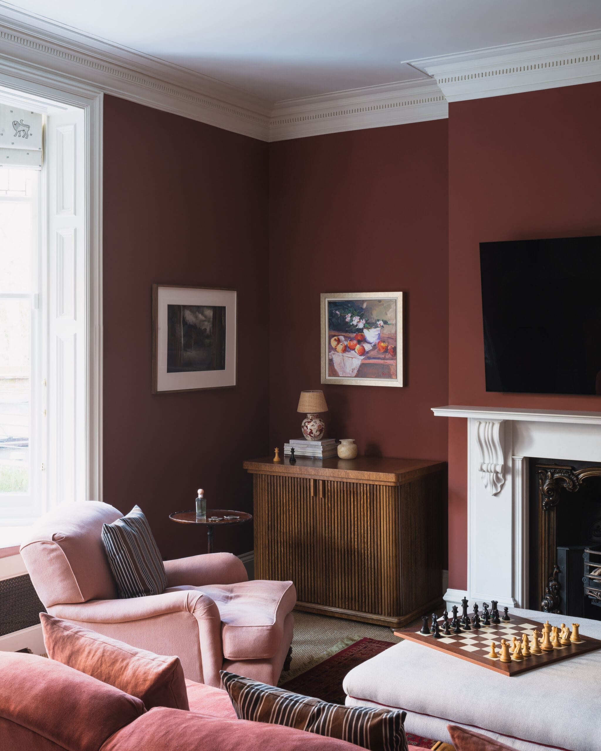 Corner of a living room with burgundy walls, white crown molding, and pink upholstered seating, plus framed art and a chessboard on a coffee table near a fireplace.”,