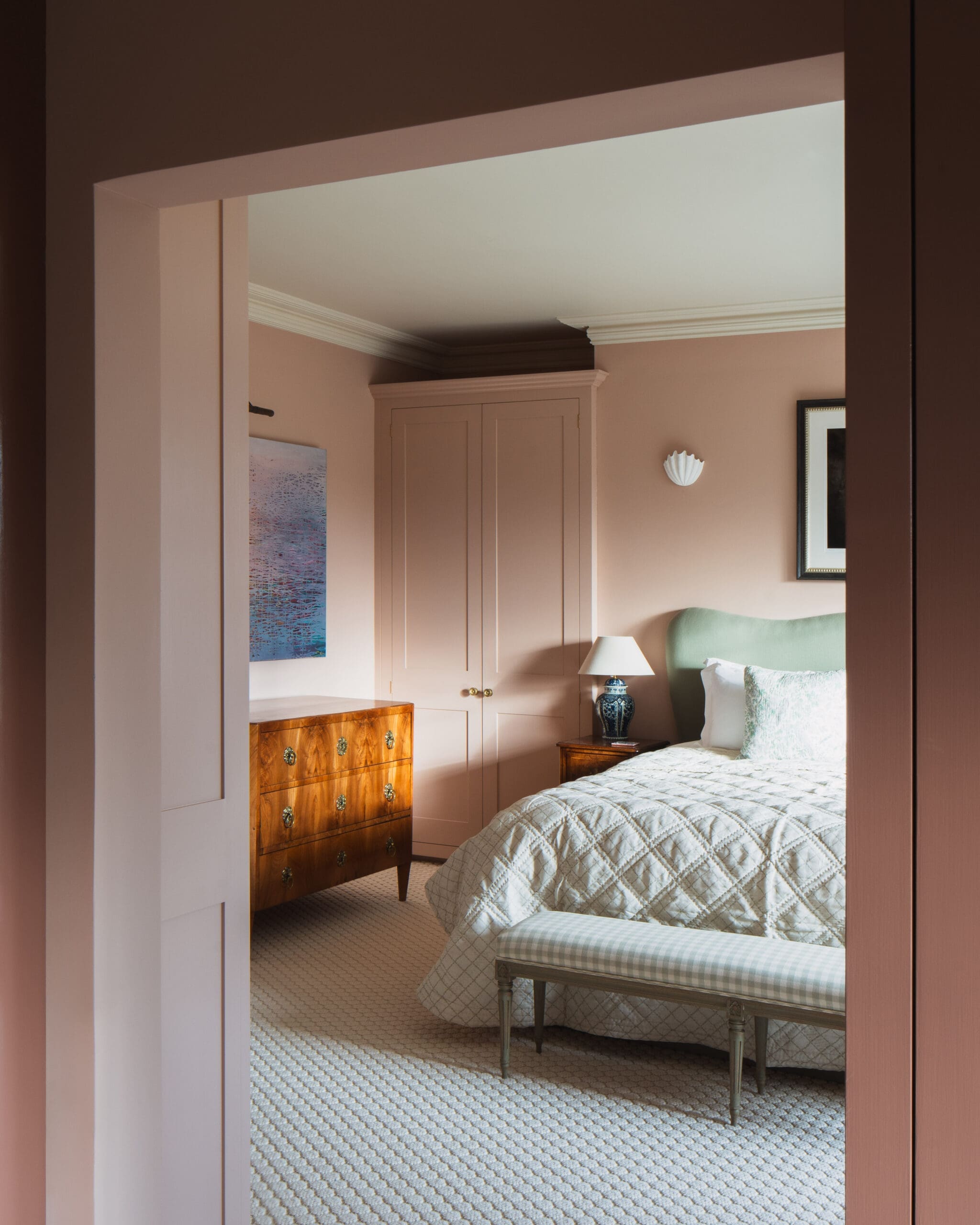 Bedroom seen through a doorway: pink walls, built-in cabinet, and a wooden dresser beside a bed with a light quilt and blue‑and‑white lamp on a side table.