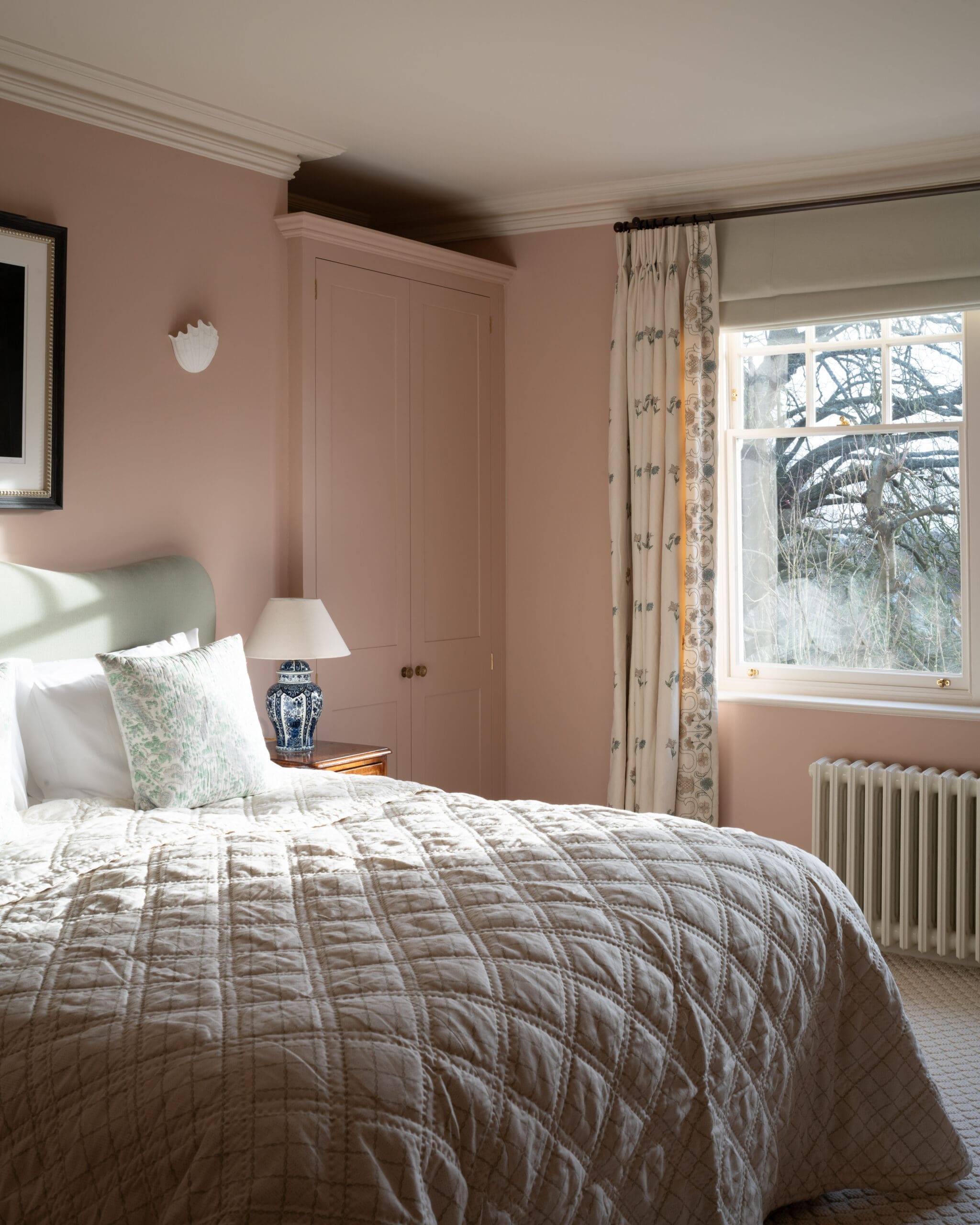 Bedroom with pale pink walls, quilted beige bedspread, and decorative pillows under a window with floral curtains.
