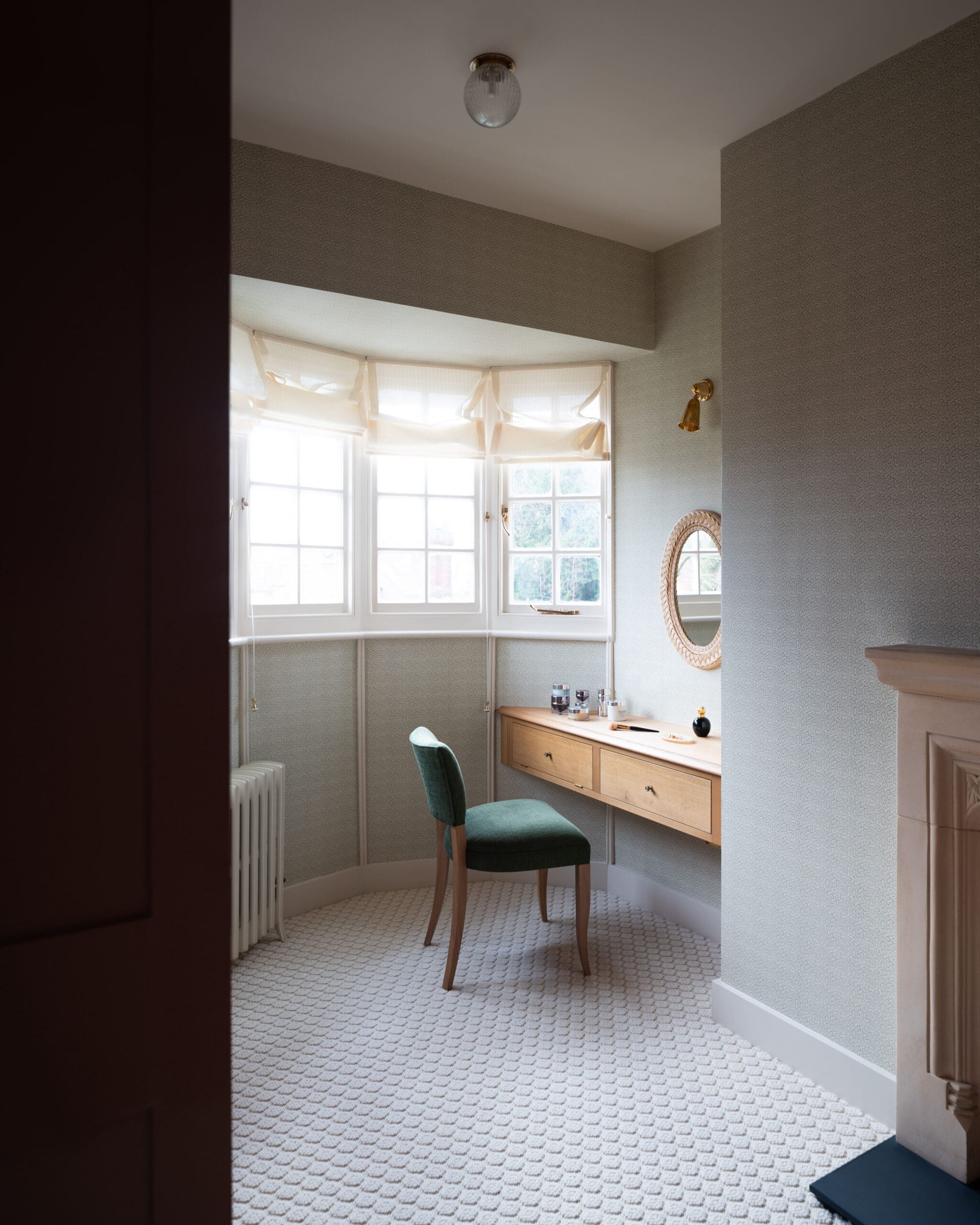 Small dressing area with a wooden desk and two drawers, a green upholstered chair, and a round wicker-framed mirror by a sunlit window with sheer curtains.