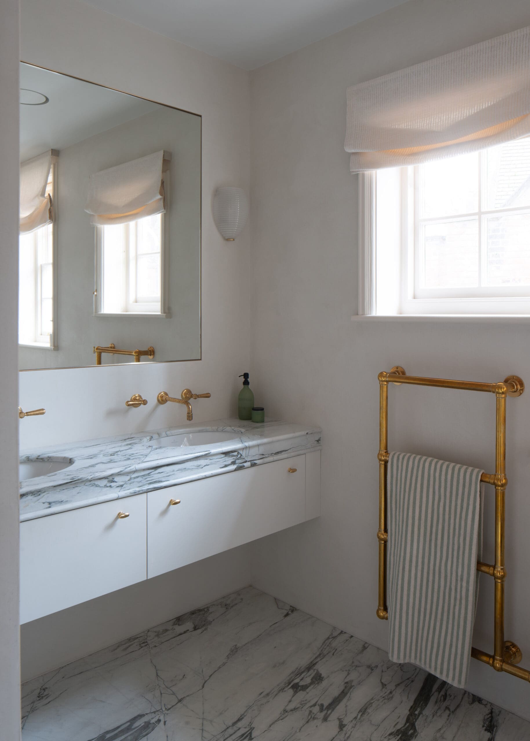 Bright bathroom with a white vanity, marble countertop, and brass fixtures beneath a large wall mirror in natural light from a window.