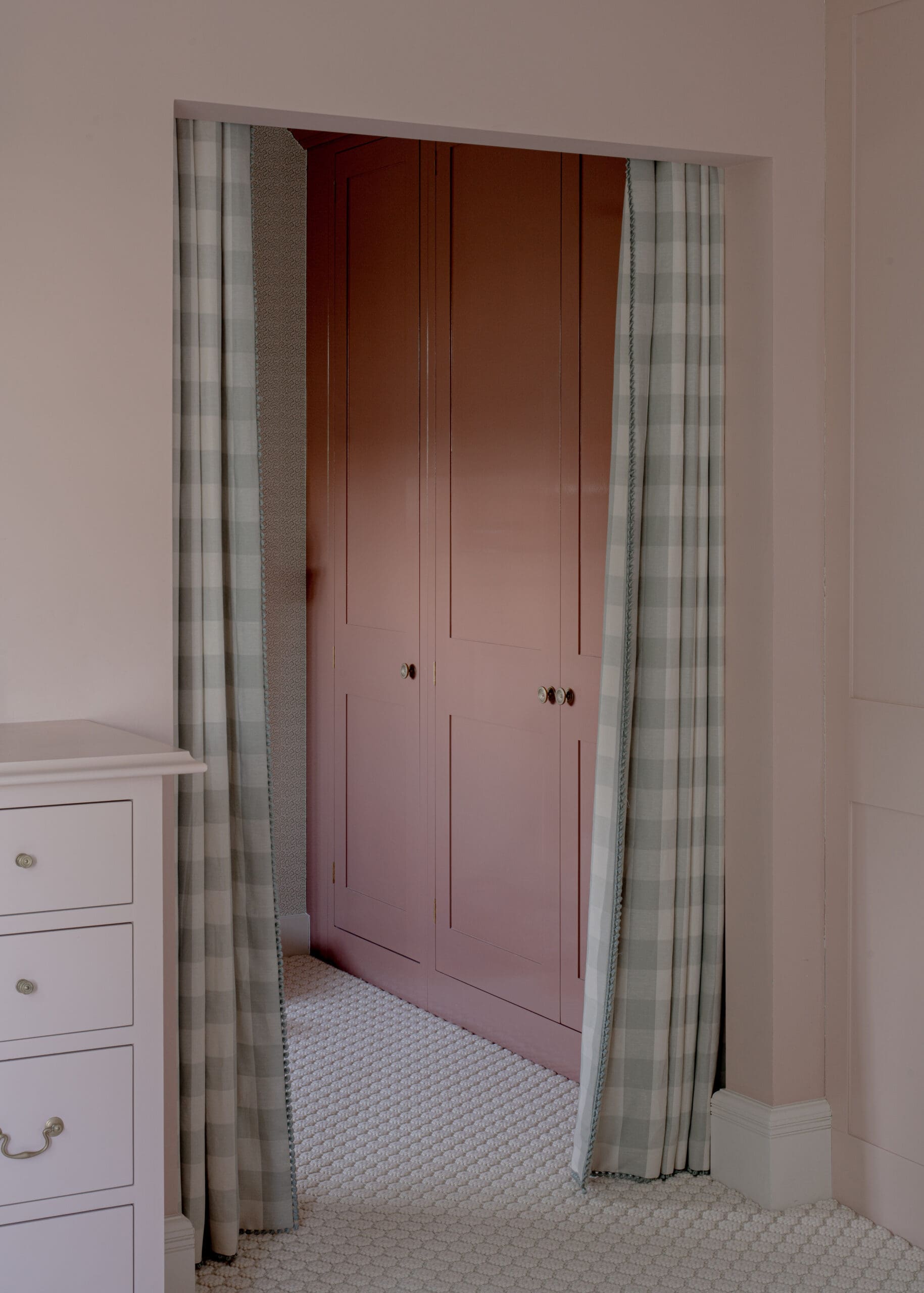 Open doorway revealing a pink built-in wardrobe with panel doors, flanked by light plaid curtains in a pastel bedroom.