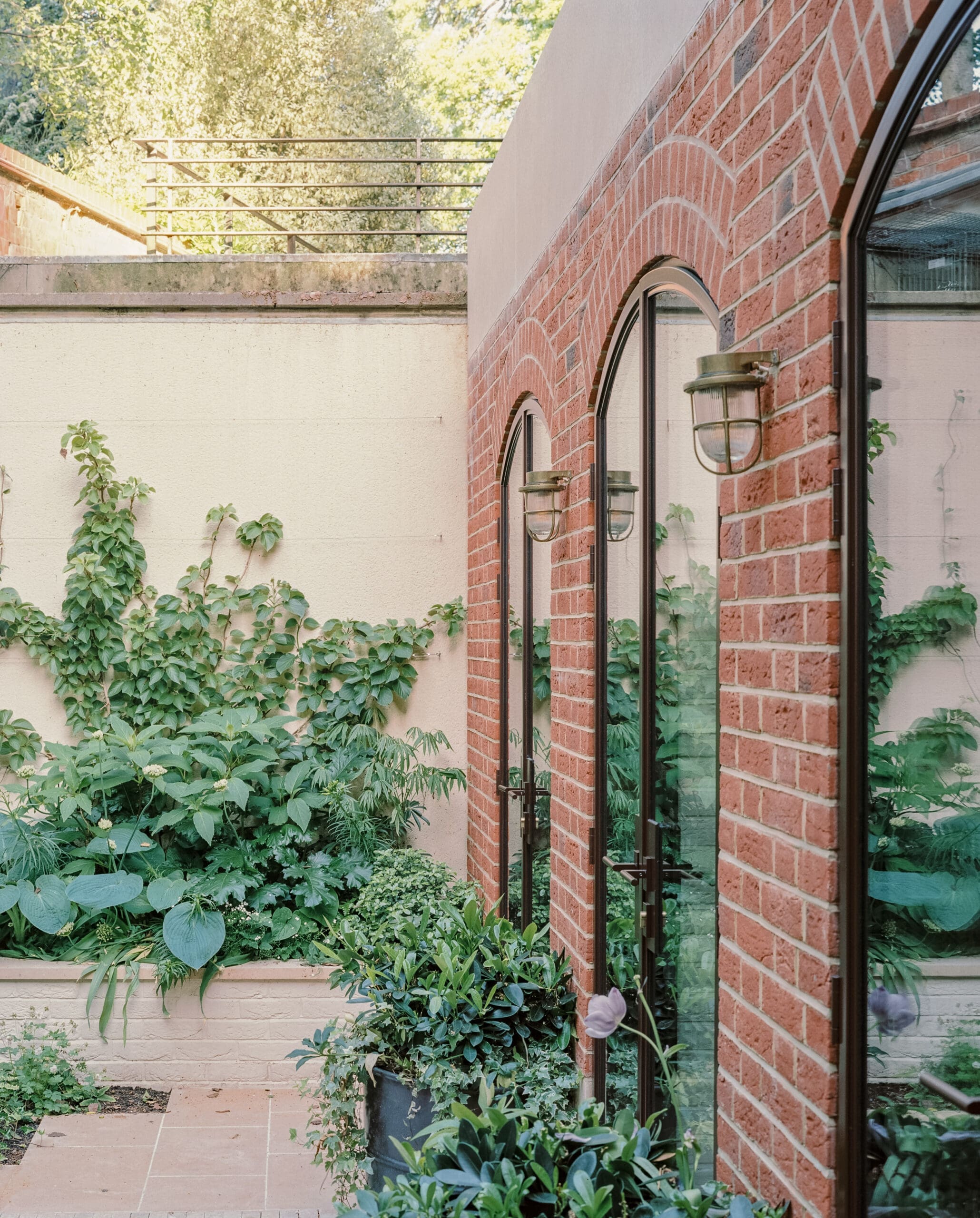 Brick wall with arched windows and outdoor lanterns, surrounded by lush green plants in a courtyard.