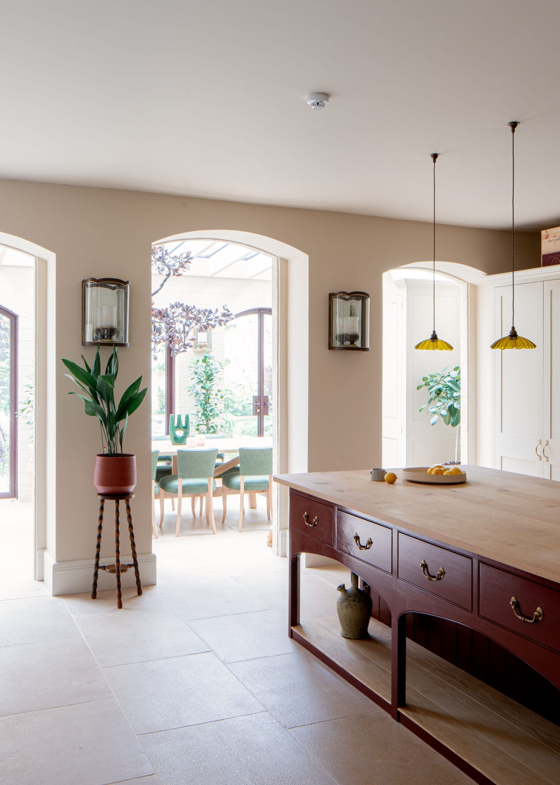 Bright kitchen/dining area with a large wooden island and hanging yellow pendant lights, arched windows, and green plants nearby.