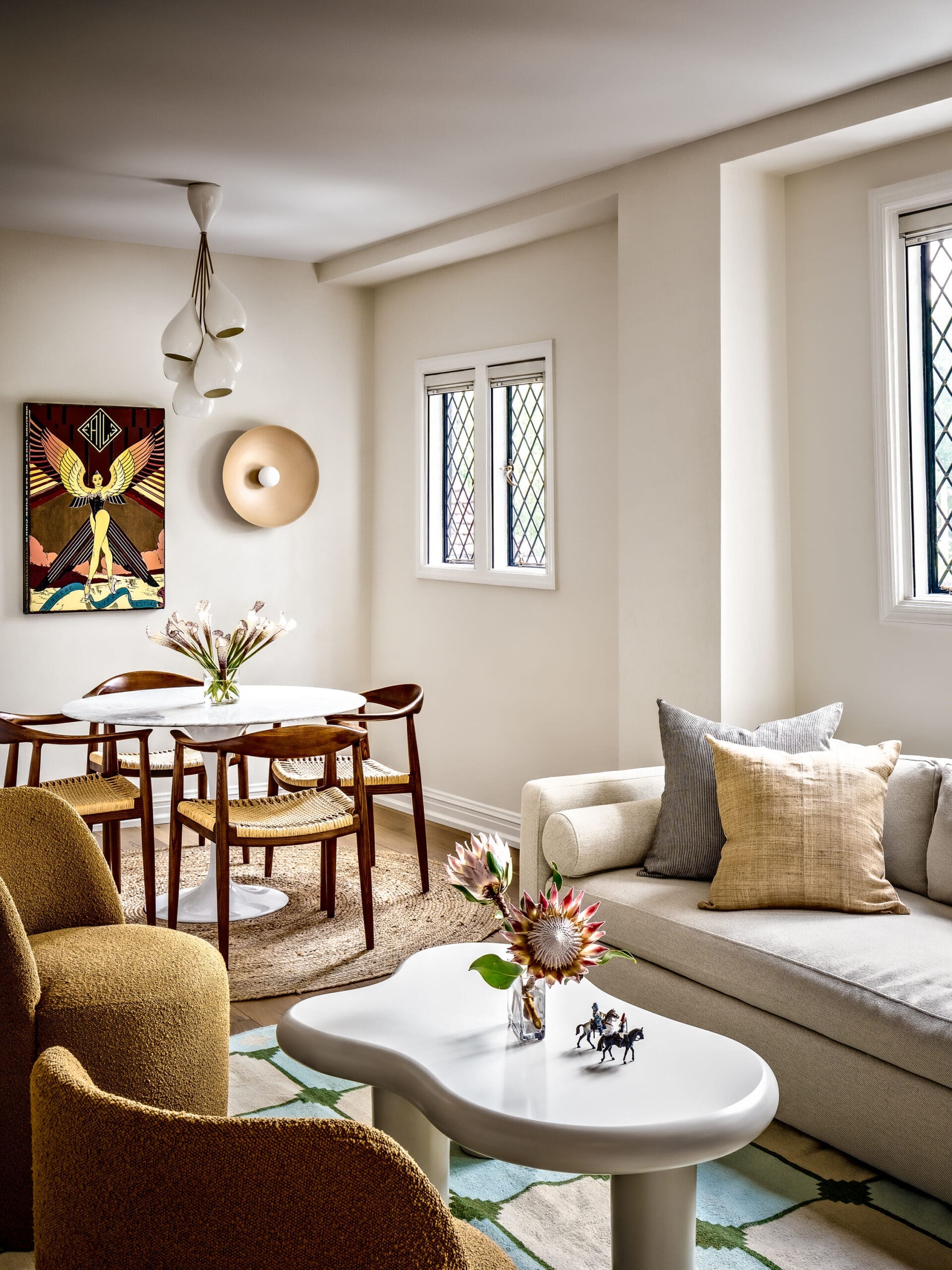Dining/living area with a white oval table, brown boucle chairs, and a beige sofa with pillows; natural light from diamond-pane windows fills the room.