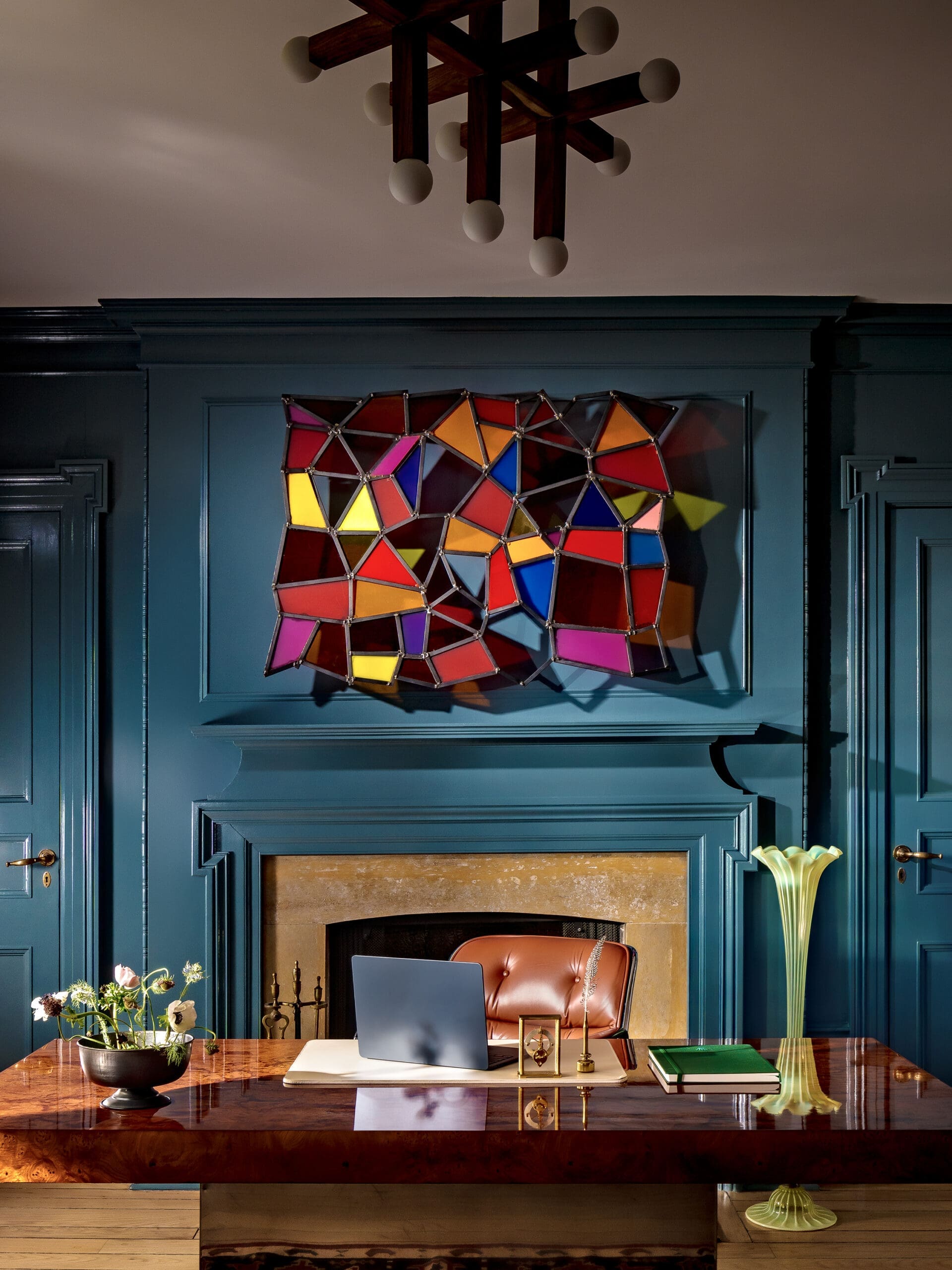 Colorful geometric stained-glass wall sculpture above a teal paneled wall, with a wooden desk and laptop in front.