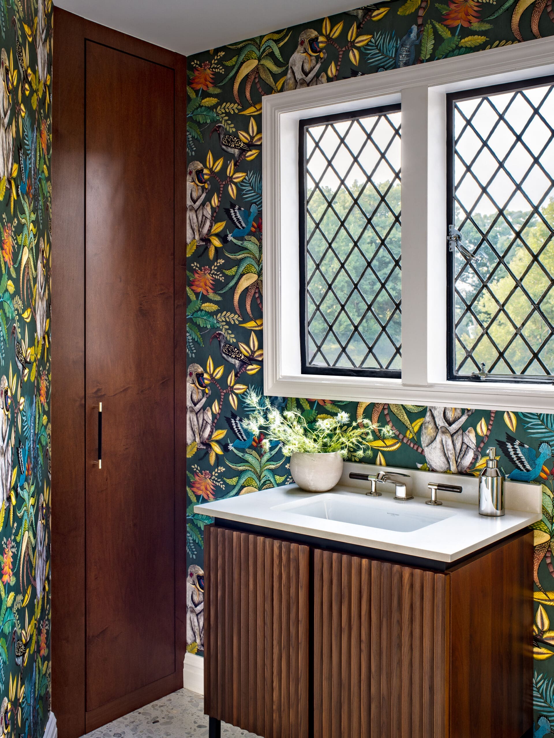 Bathroom with dark jungle wallpaper, a tall wooden cabinet, white sink with chrome faucet, and a diamond-pattern window.