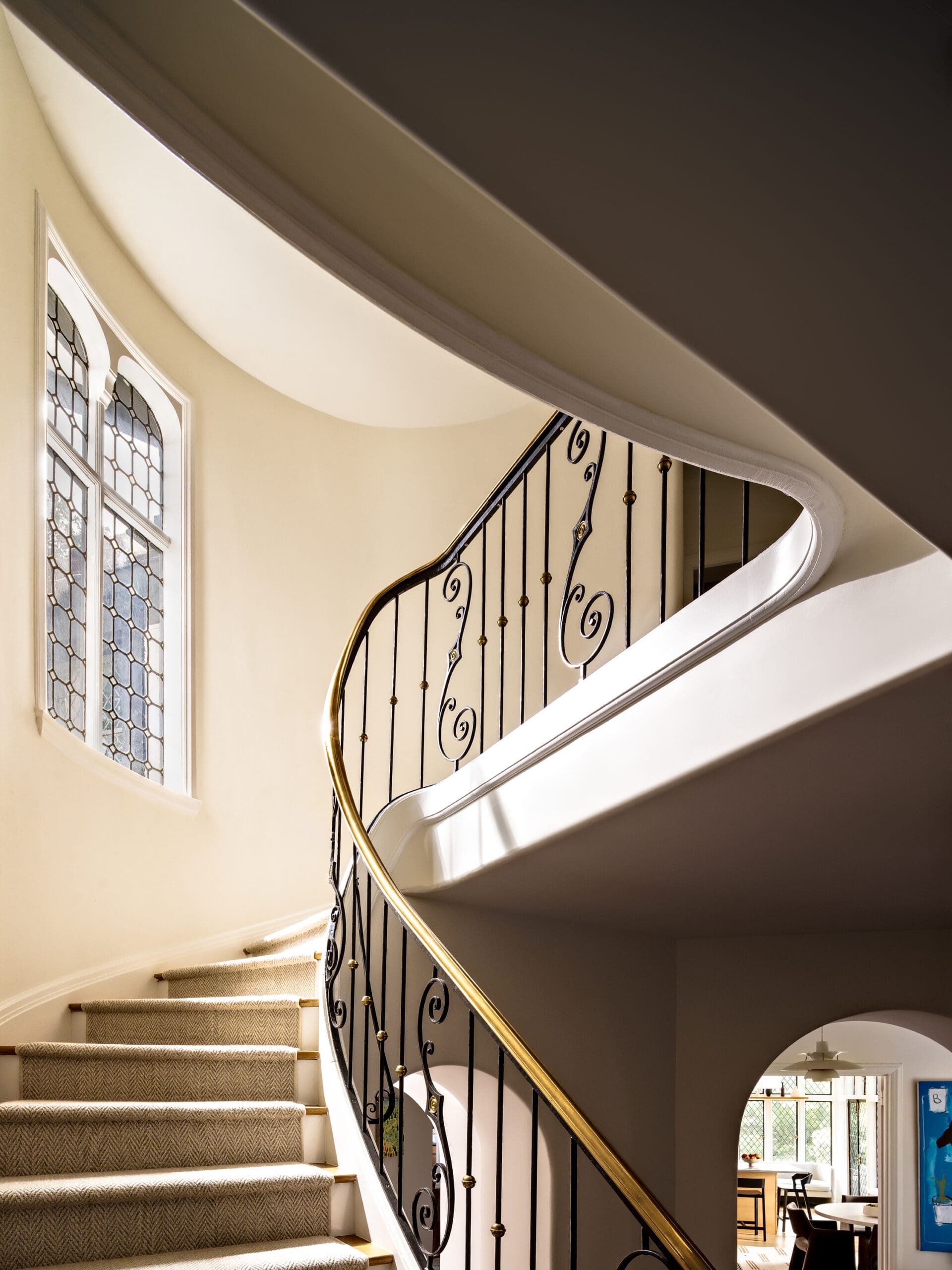 Curved stairway with black iron railing and gold handrail, beige walls, and a stained-glass window on the left.