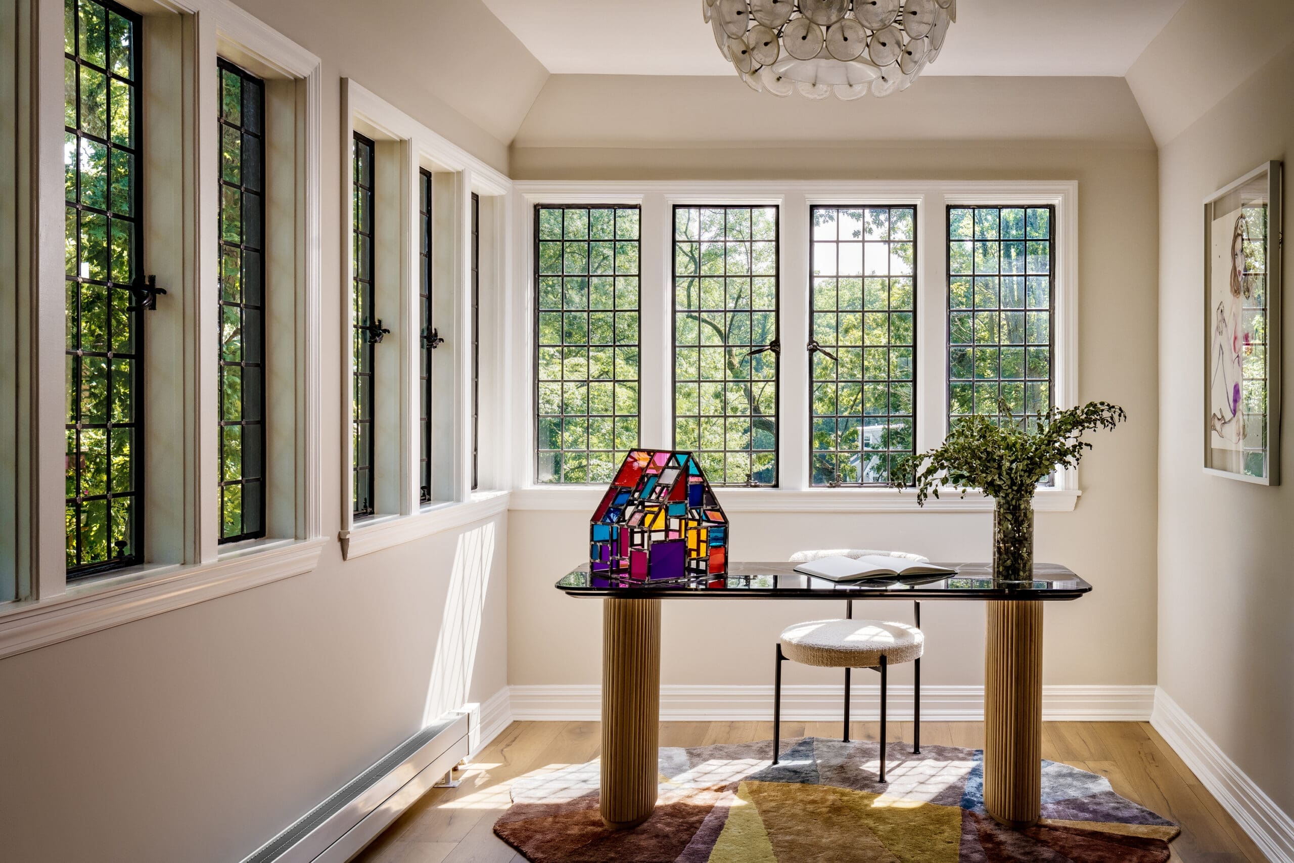 Bright corner office with tall grid windows, a glass pedestal table, and a colorful stained-glass sculpture centerpiece. A potted plant and notebook sit nearby.
