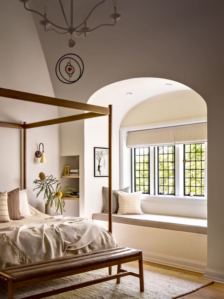 Bedroom with a wooden canopy bed, beige bedding, and a window seat beside a large arched window.