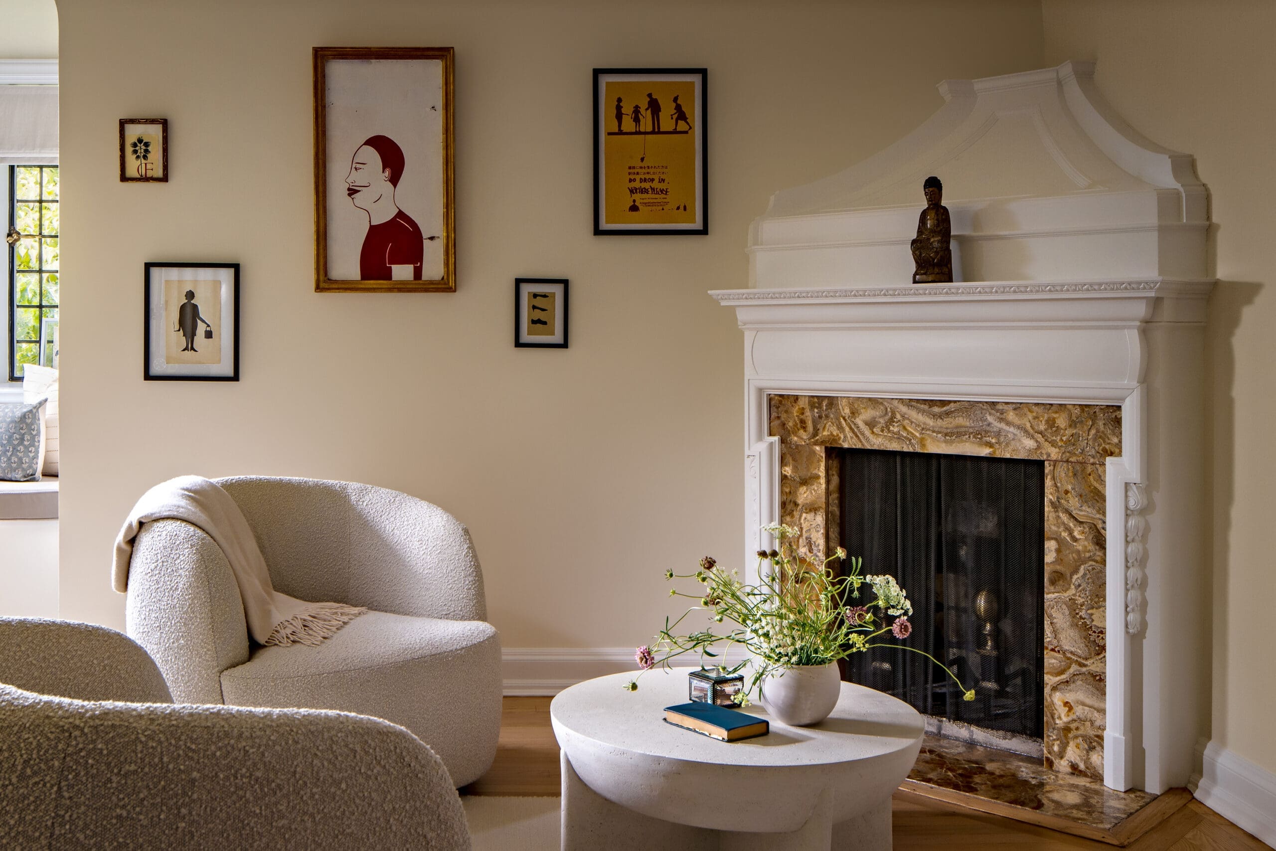 Cozy living room with a white ornate fireplace, beige walls, and framed art; a round coffee table with flowers and a couple of books sits in front of a beige sofa.