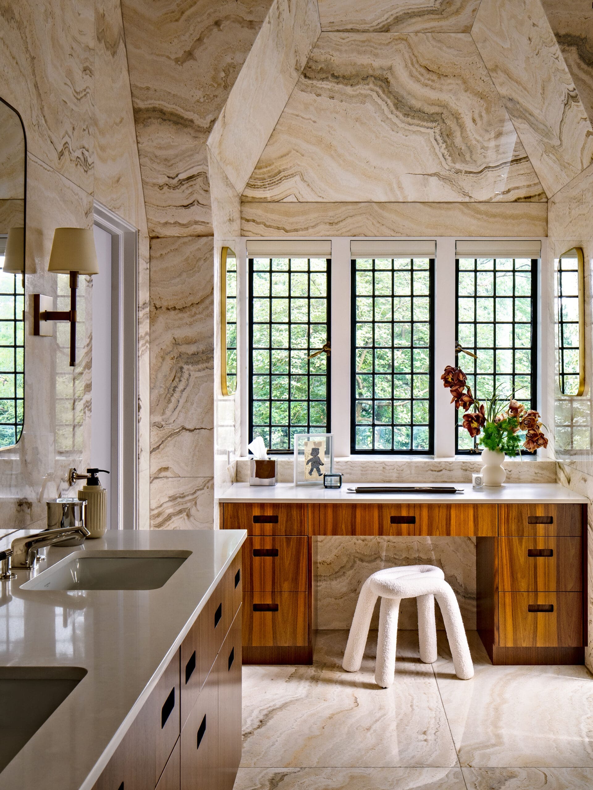 Modern kitchen with beige marble walls and wooden cabinets, white countertop, and a white stool by the windowed wall.