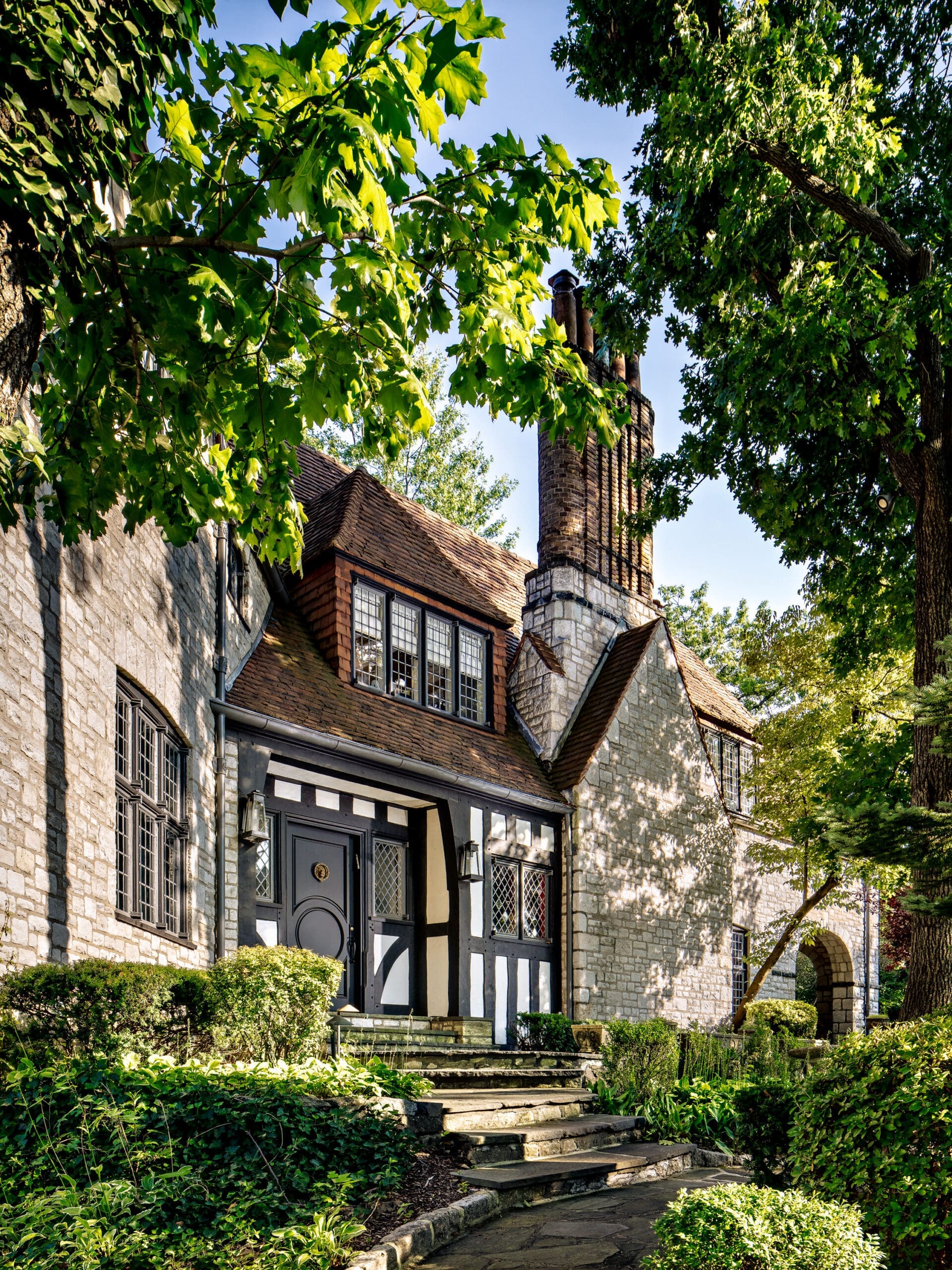 Historic stone cottage with black-and-white timber framing, ivy and lush garden, stone steps to a dark front door beneath leafy trees.