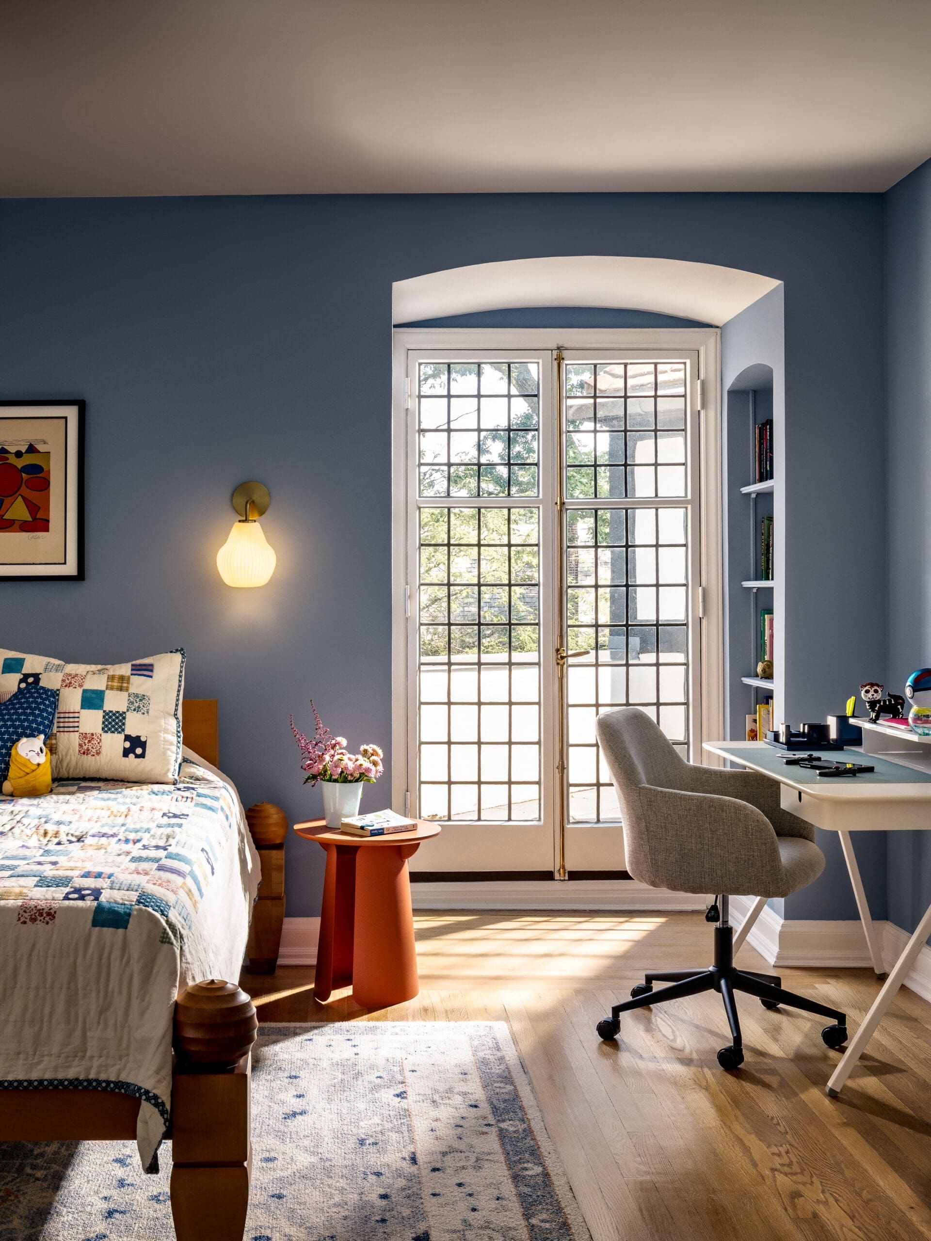 Blue bedroom with a patchwork quilted bed, orange side table, and a potted plant beside tall French doors that let in sunlight.