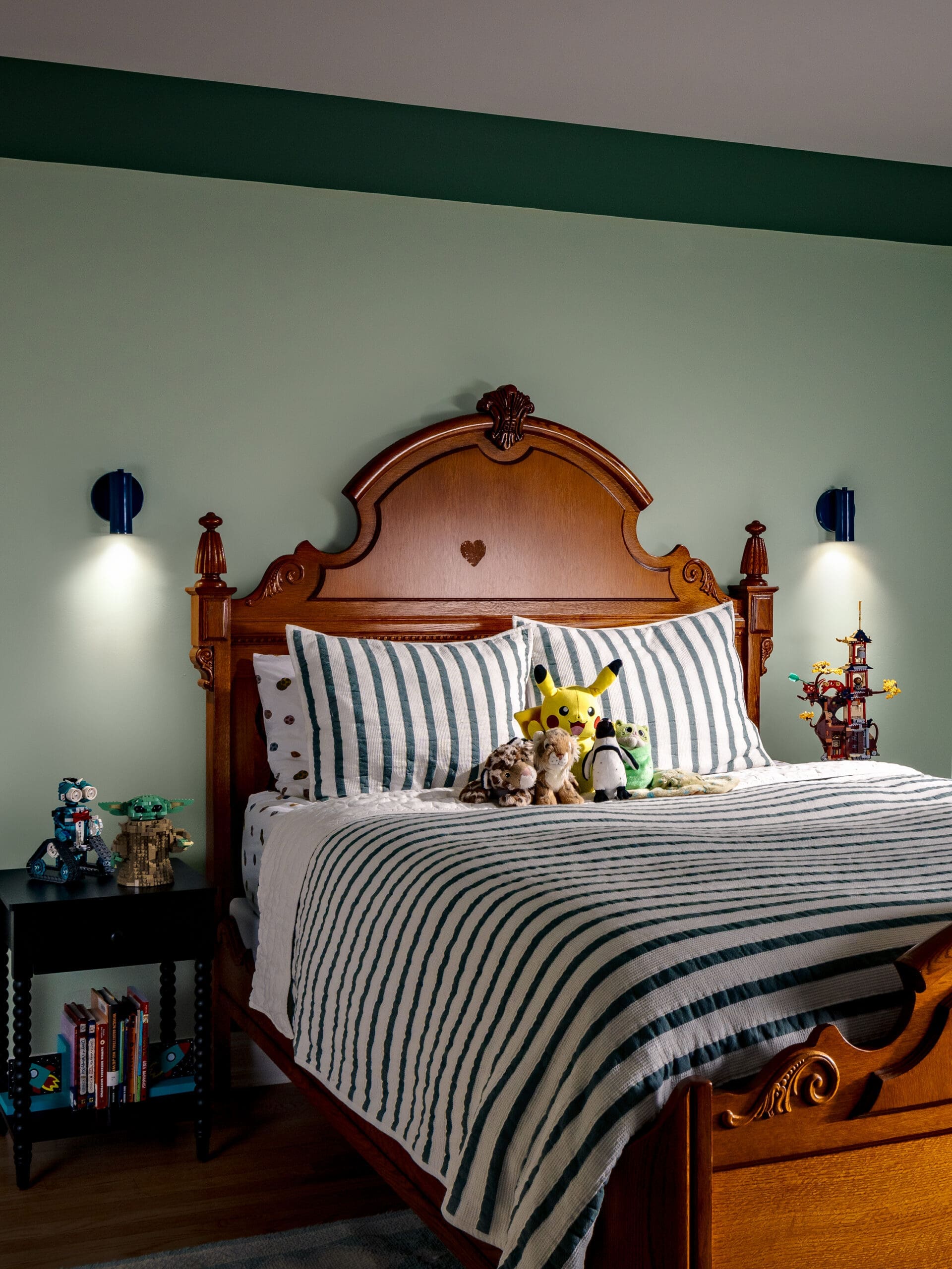 Child's bedroom with a carved wooden bed, green walls, and striped bedding; plush animals lined on the bed and a bookshelf with LEGO figures beside it.
