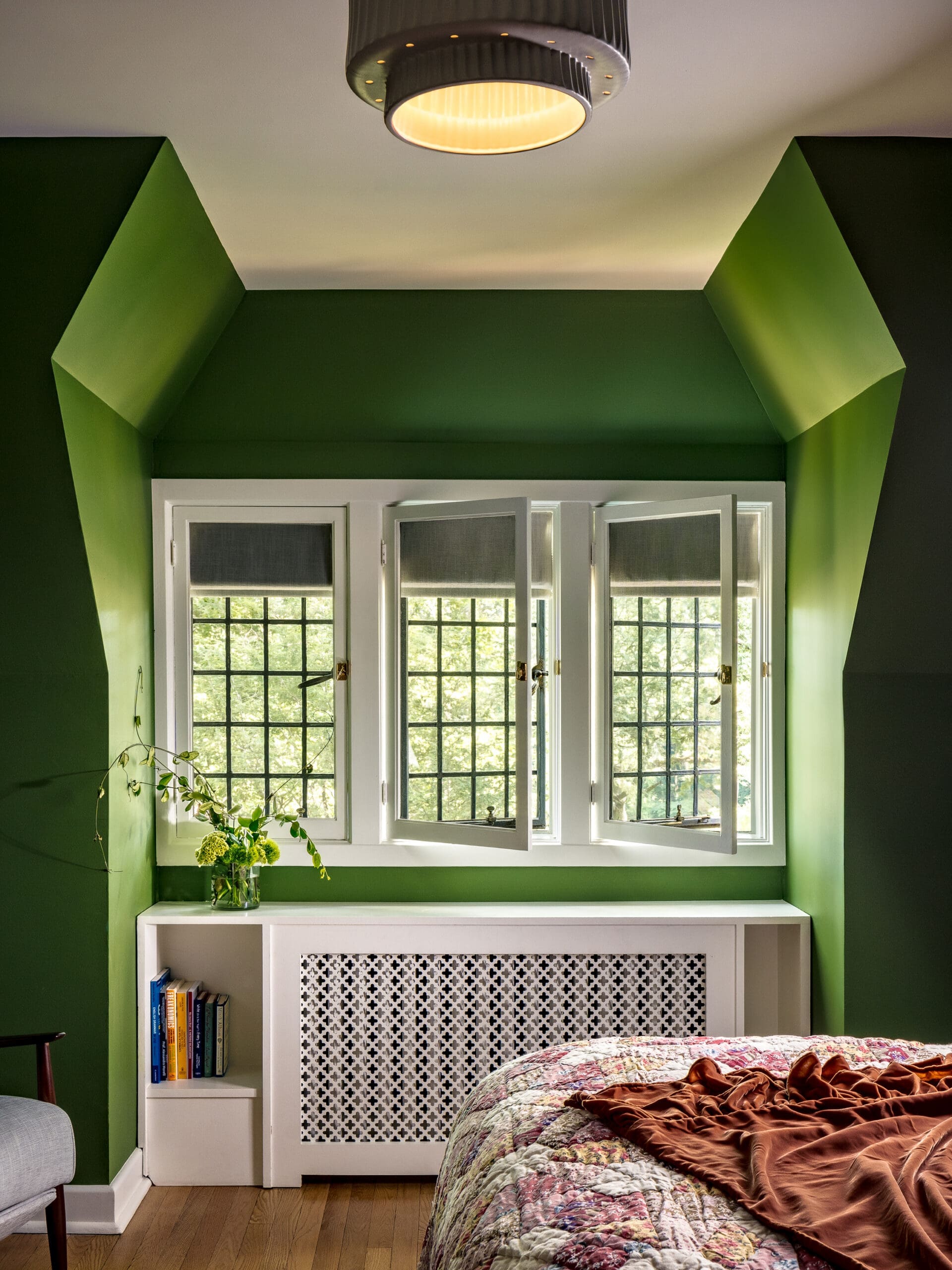 Green-walled bedroom with a white window seat and radiator cover featuring a lattice panel, a small bookshelf, and a quilted bed with brown blanket.
