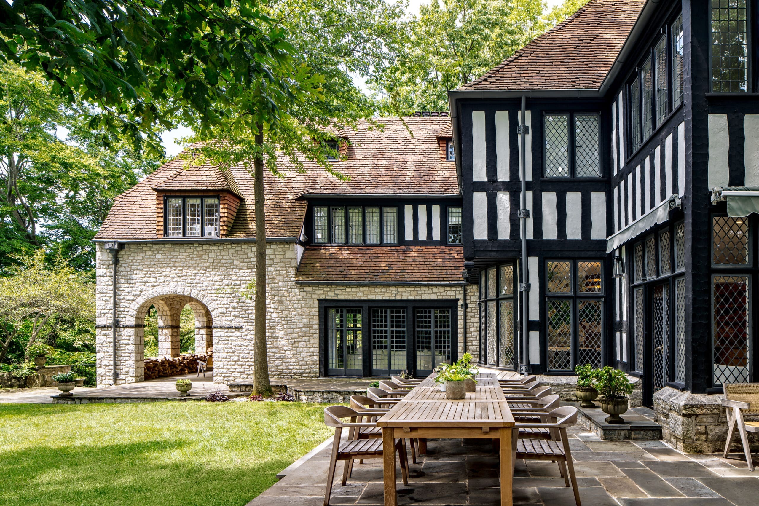 Courtyard scene with a stone-and-timbered building, stone arches, and long wooden tables under a leafy canopy.