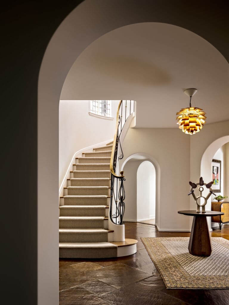 Elegant curved staircase with carpeted steps, ornate black railing, and warm hanging lamp in a spacious foyer.