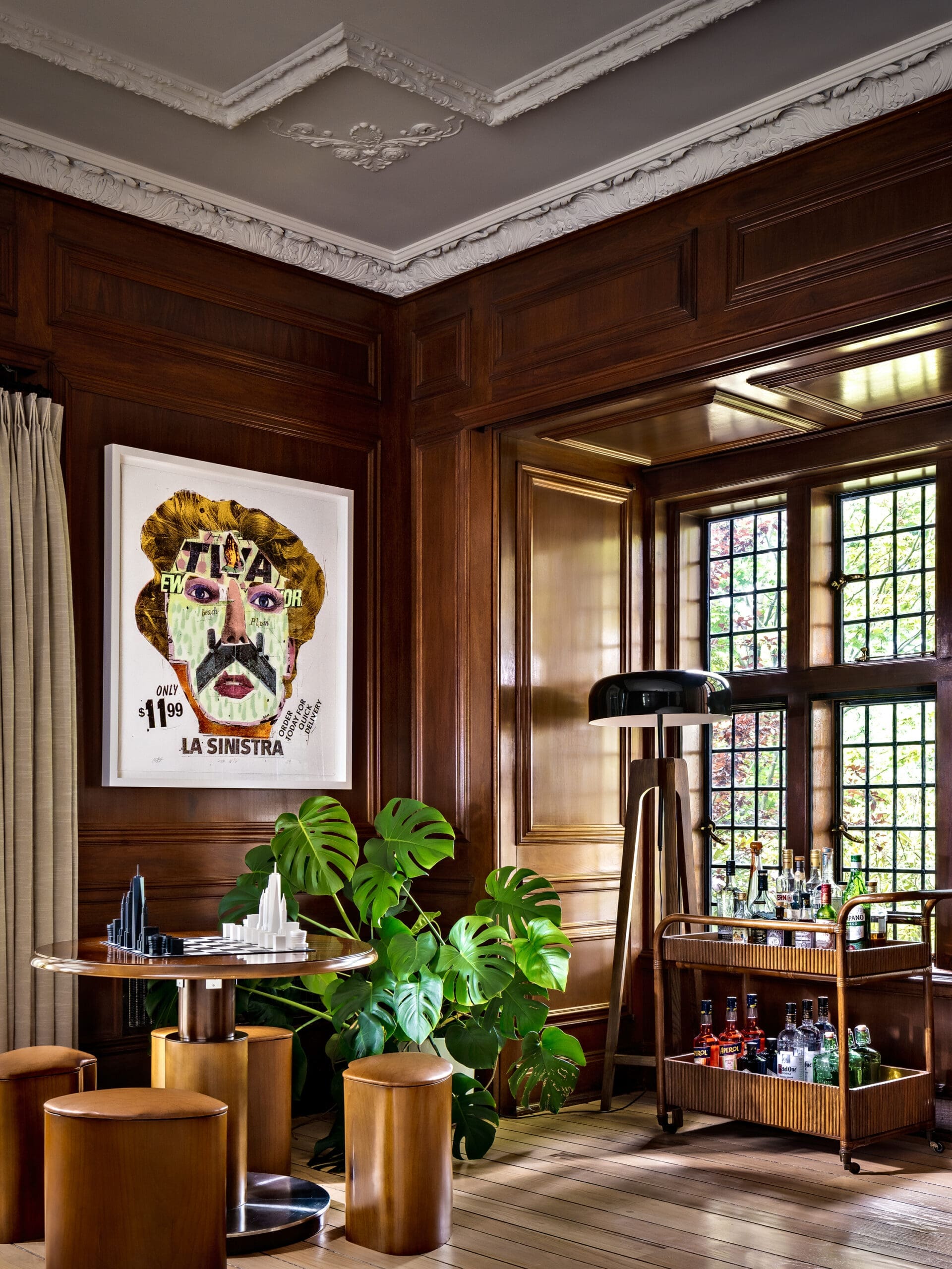 Wood-paneled room with a framed colorful portrait, lush green plants, and a round-topped table by a windowed bar area.
