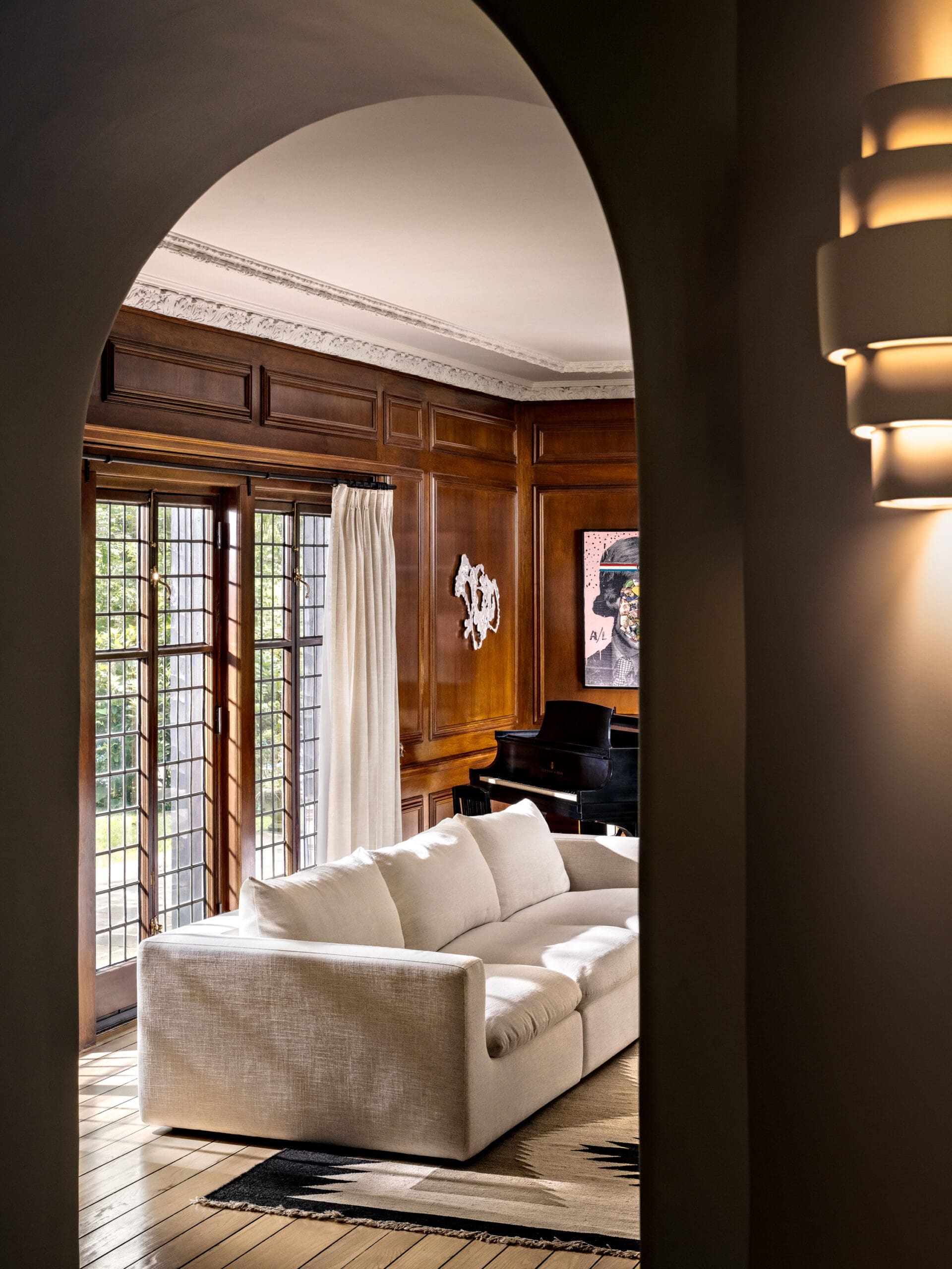 Archway frames a traditional living room with dark wood paneling, a beige sofa, and a grand piano near grid-pattern windows.