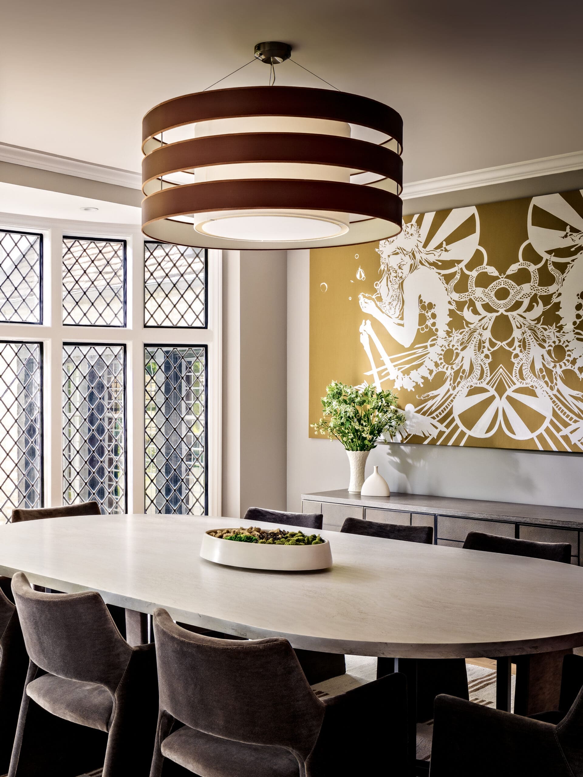 Round, multi-ring chandelier over a light dining table in a contemporary room with dark chairs and a gold wall mural.
