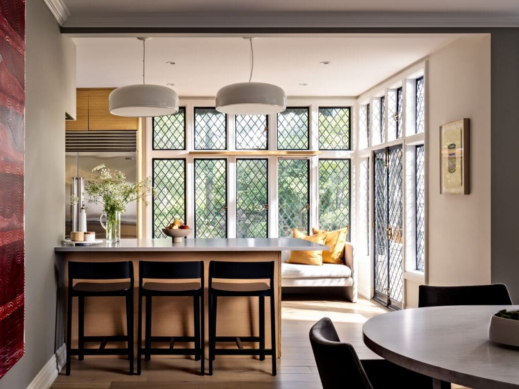 Kitchen island with black stools, a fruit bowl, and tall diamond-pane windows providing natural light on a warm, modern space with pendant lamps above the island.