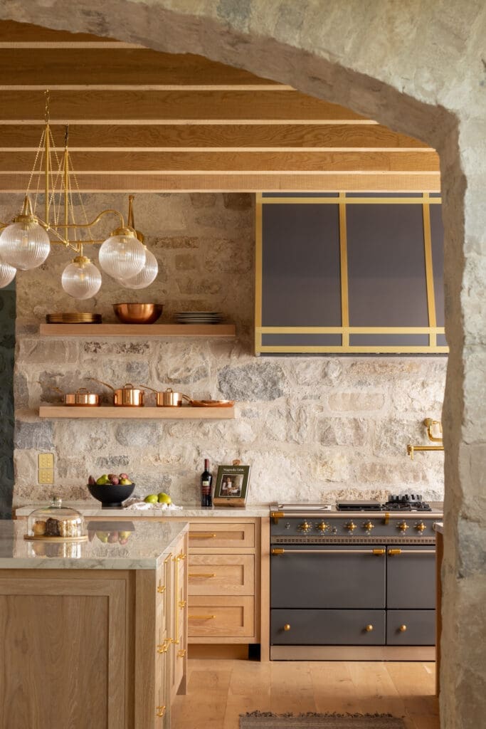 Rustic stone-kitchen with wooden cabinets, copper pots on a shelf, and a brass-trimmed range.