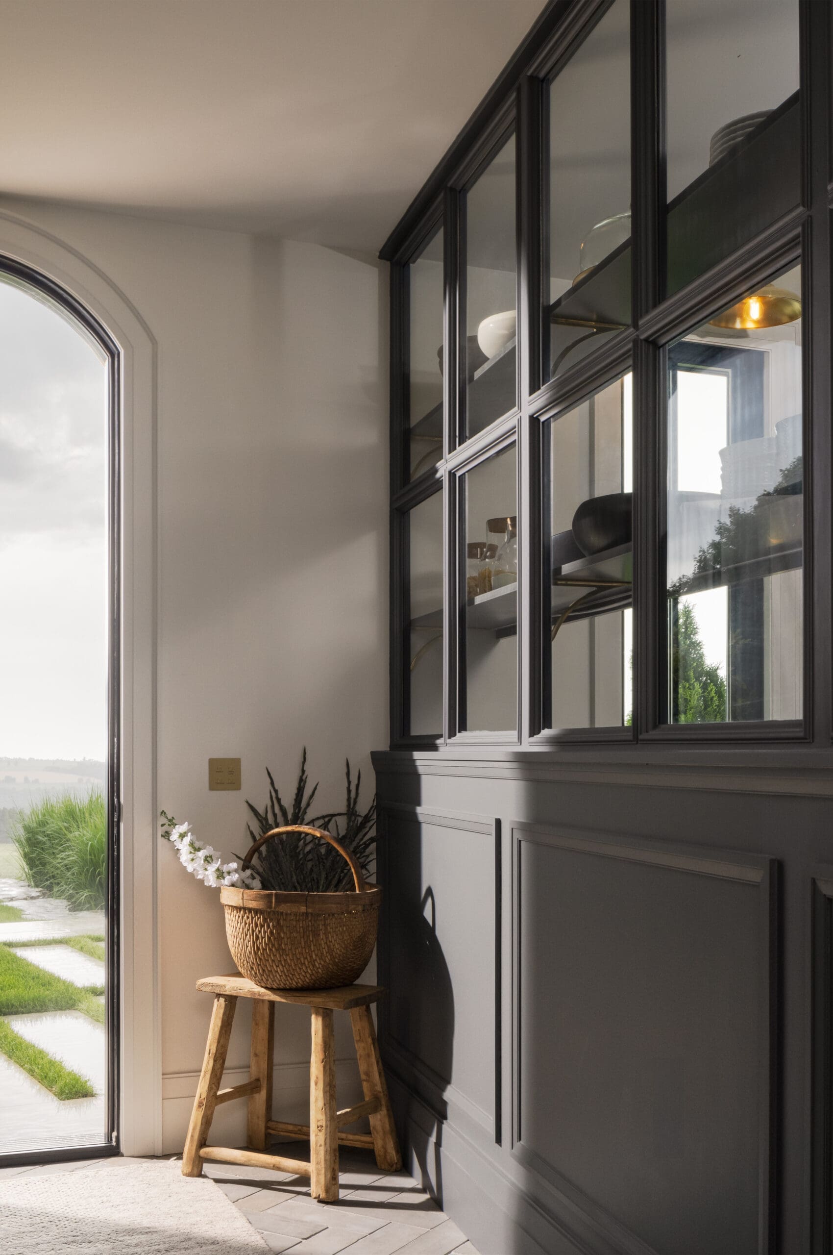 Dark-framed glass-front cabinets run along a gray wall beside a light entry with an arched doorway and a wicker basket of white flowers on a wooden stool.
