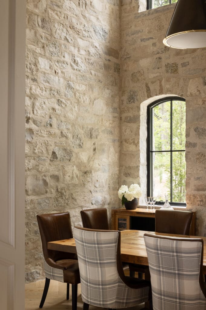 Stone-walled dining area with a wooden table, brown leather chairs, and a window showing greenery outside.
