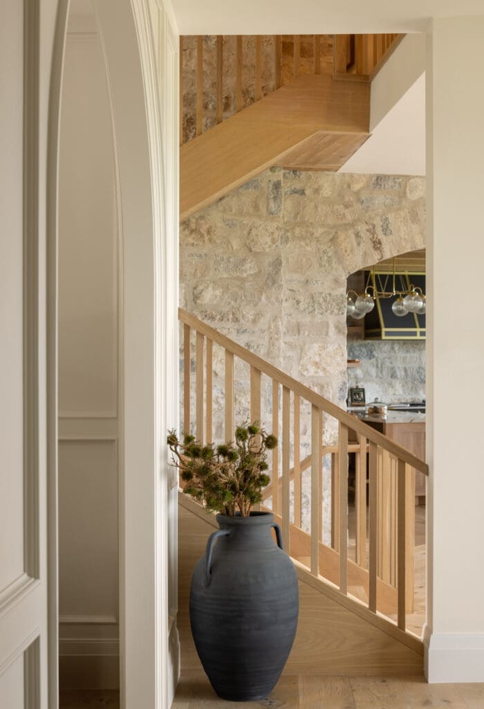 Wood and stone stair hall with a curved wooden railing, exposed stone wall, and a large dark ceramic vase with greenery in the foreground