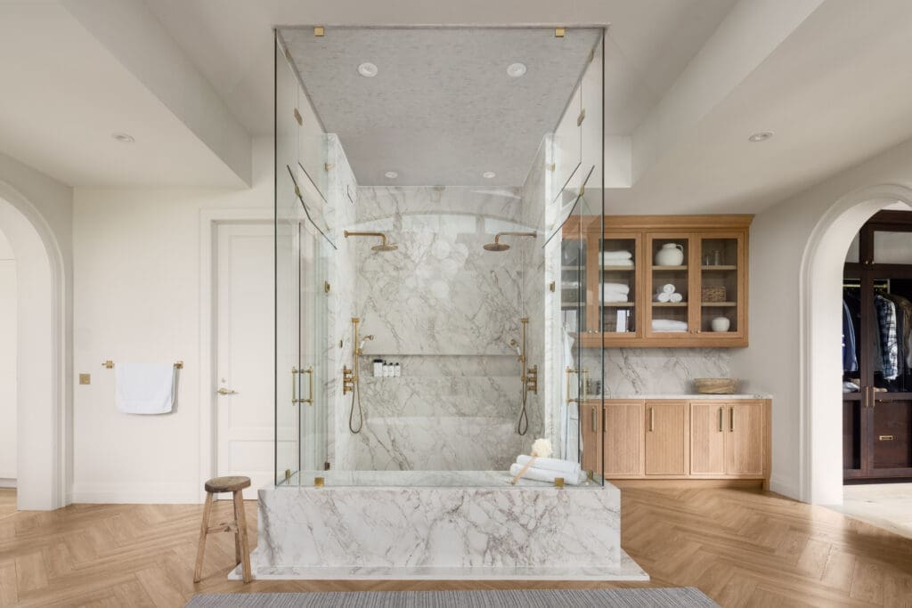 Glass-walled walk-in shower with marble walls and brass fixtures, flanked by a wooden cabinet and open closet area.