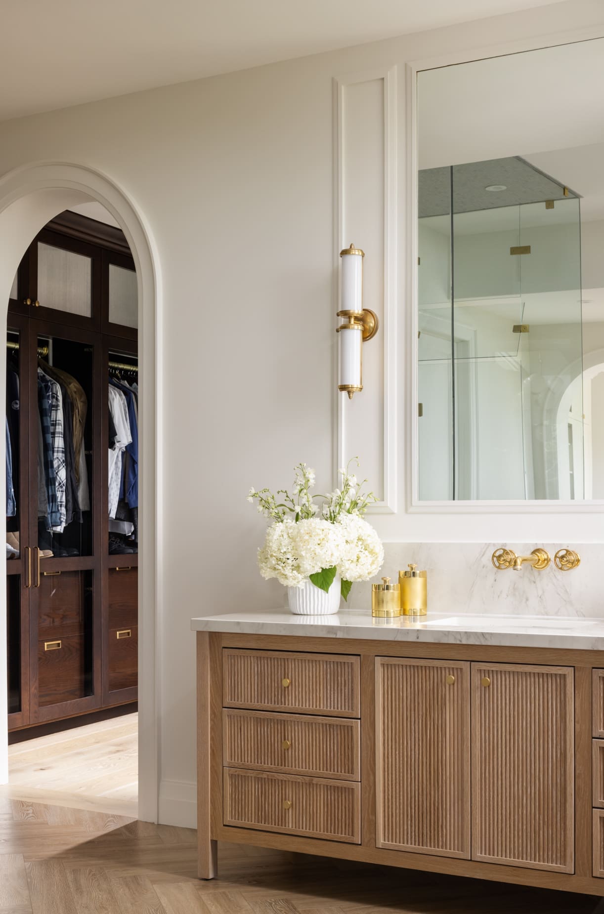 Bathroom vanity with marble top, gold faucet and hardware, and a vase of white hydrangeas; arched doorway to a closet with hanging clothes.