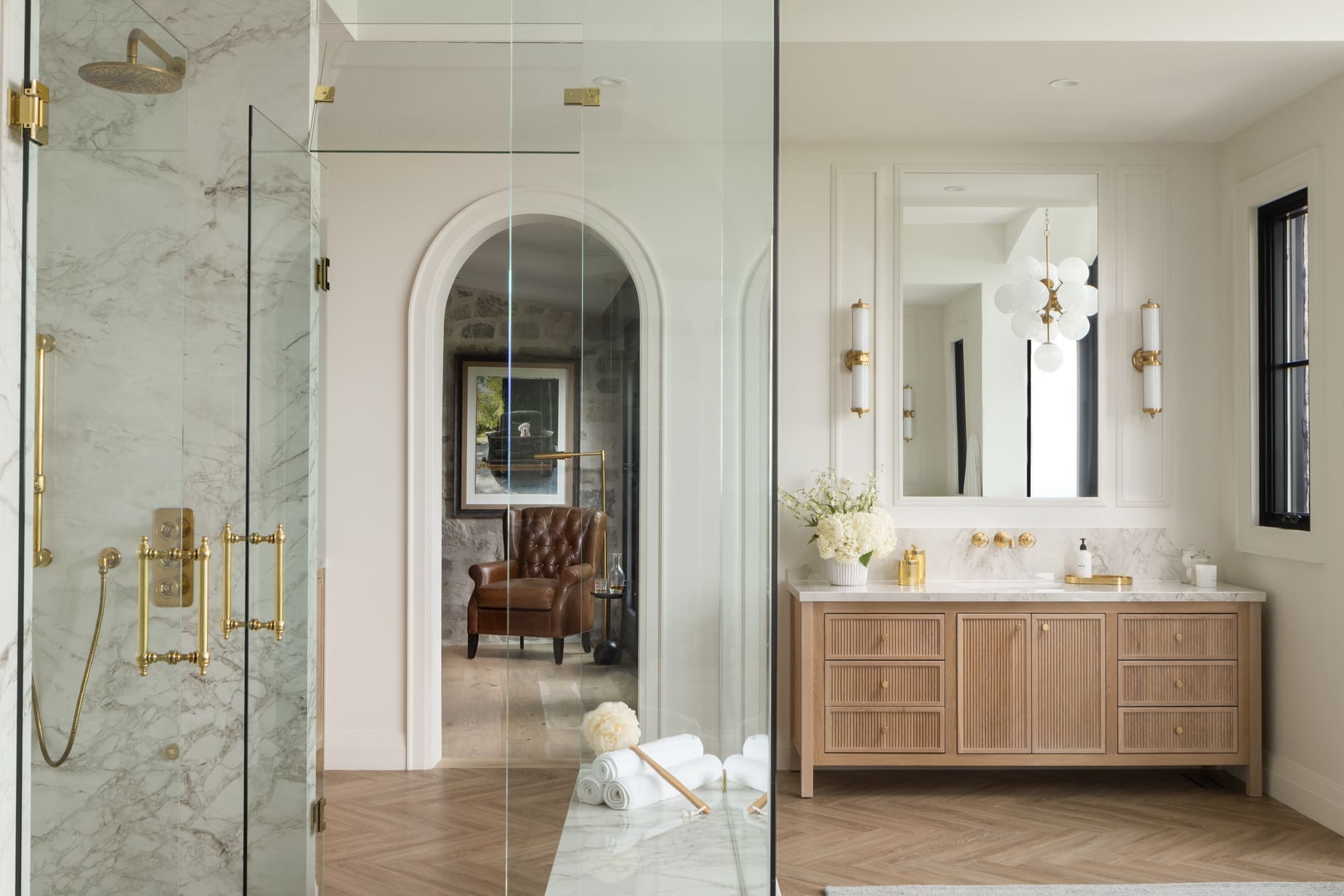 Luxurious bathroom with a glass shower enclosure, marble walls, and brass fixtures paired with a wooden vanity and large mirror.