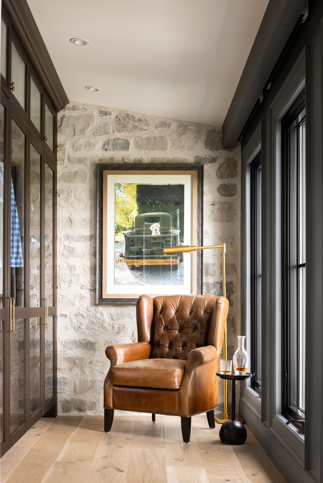 Leather brown tufted armchair in a stone-walled corner, with a small round table and decanter nearby; framed photo of a truck outside hangs on the wall behind.